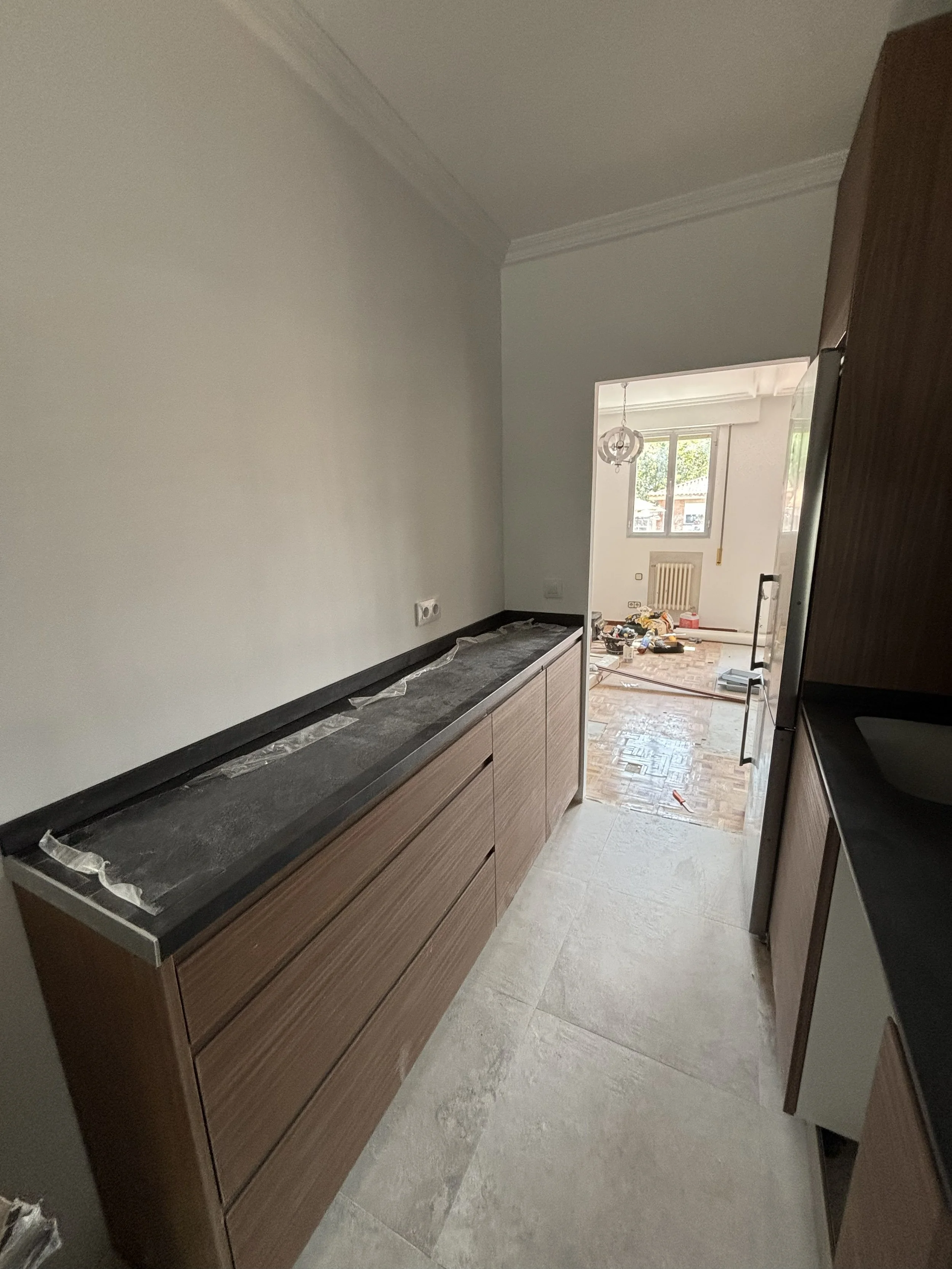 Cocina en construcción con gabinetes de madera y encimera de piedra en proceso de instalación, vista desde la entrada hacia una habitación con ventanas y piso de parquet.