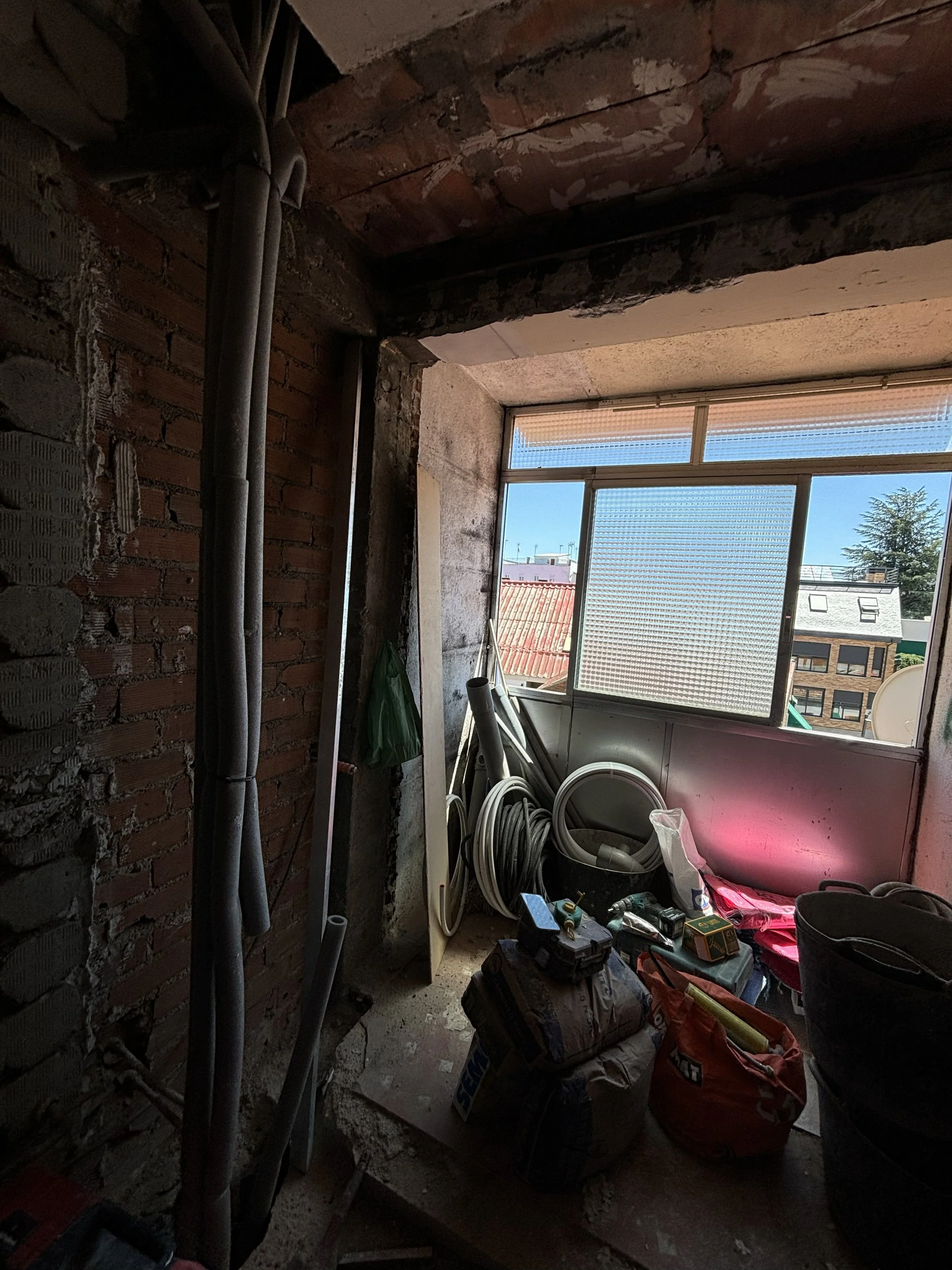 Espacio en construcción o renovación con herramientas, tubos y materiales junto a una ventana con vistas a edificios y un cielo azul.