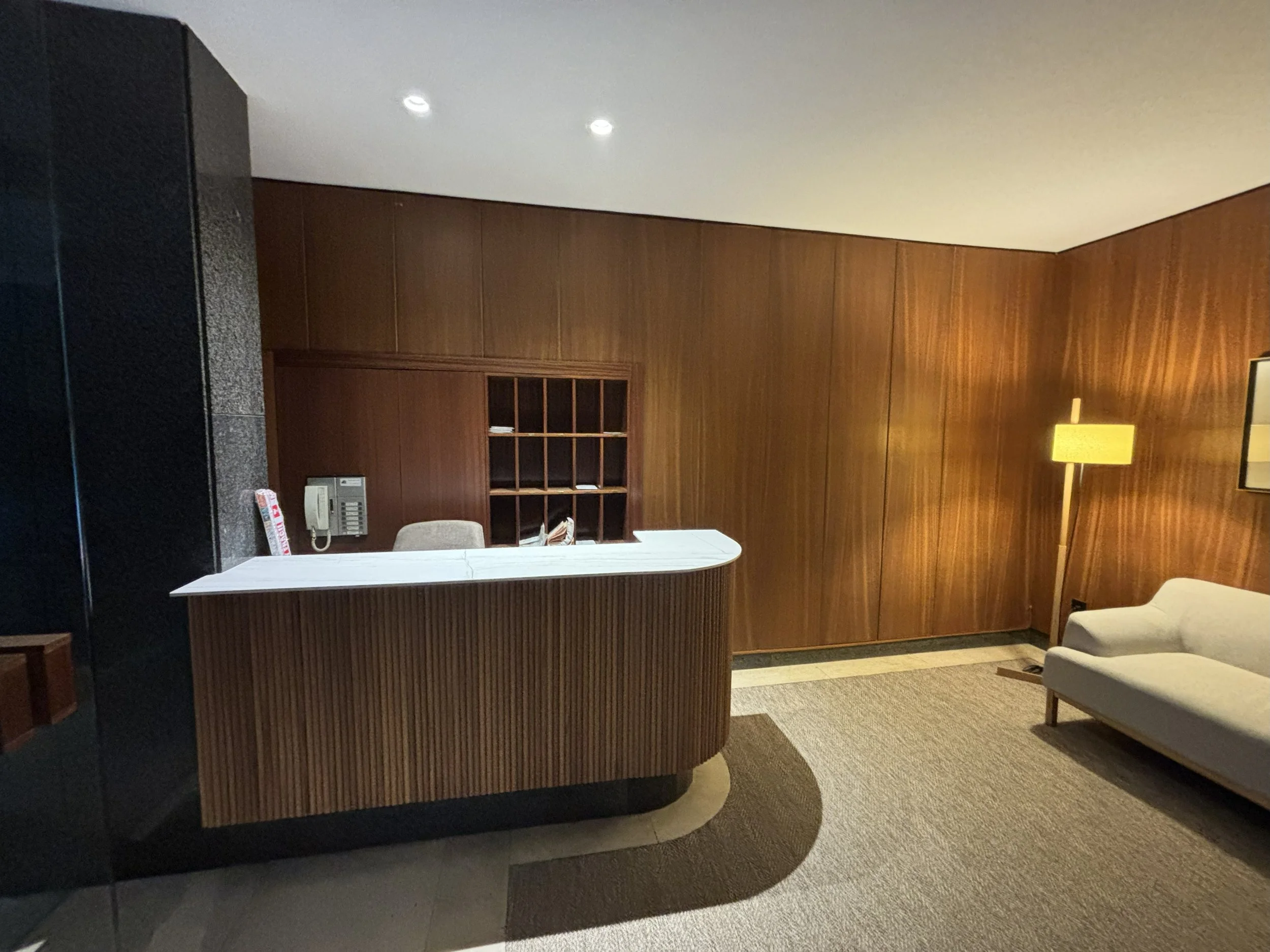 Recepción de una oficina o hotel con escritorio de madera, silla, teléfono y estantes con documentos, pared de madera, lámpara y sillón beige.