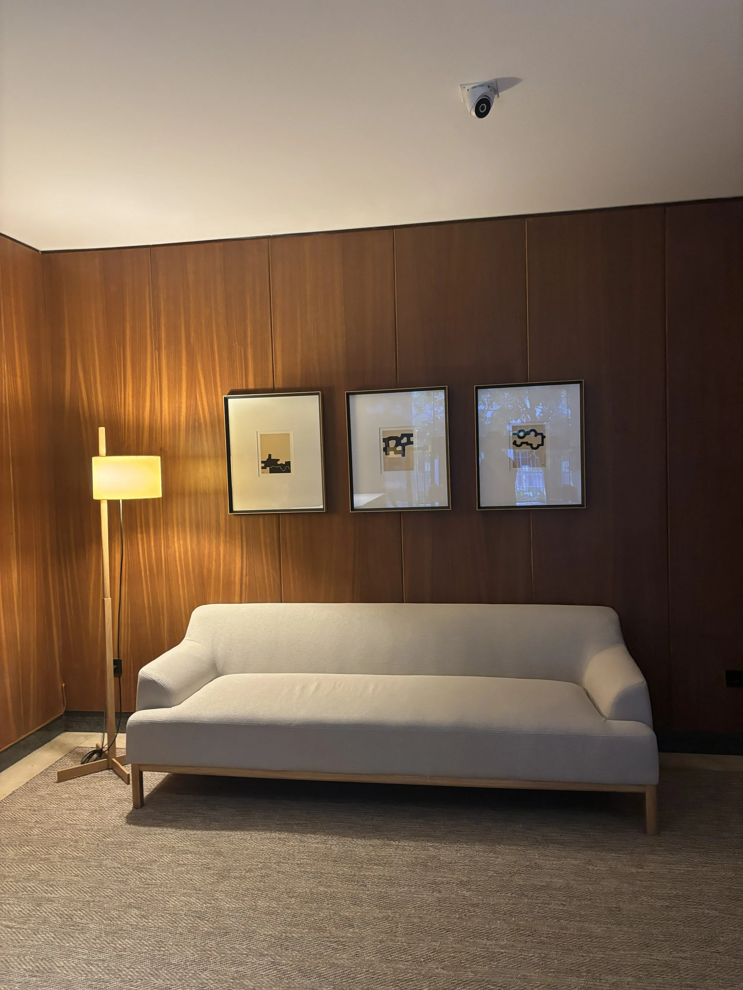 Sala de estar con pared de madera, sillón blanco, lámpara de pie y tres cuadros con arte abstracto en la pared.