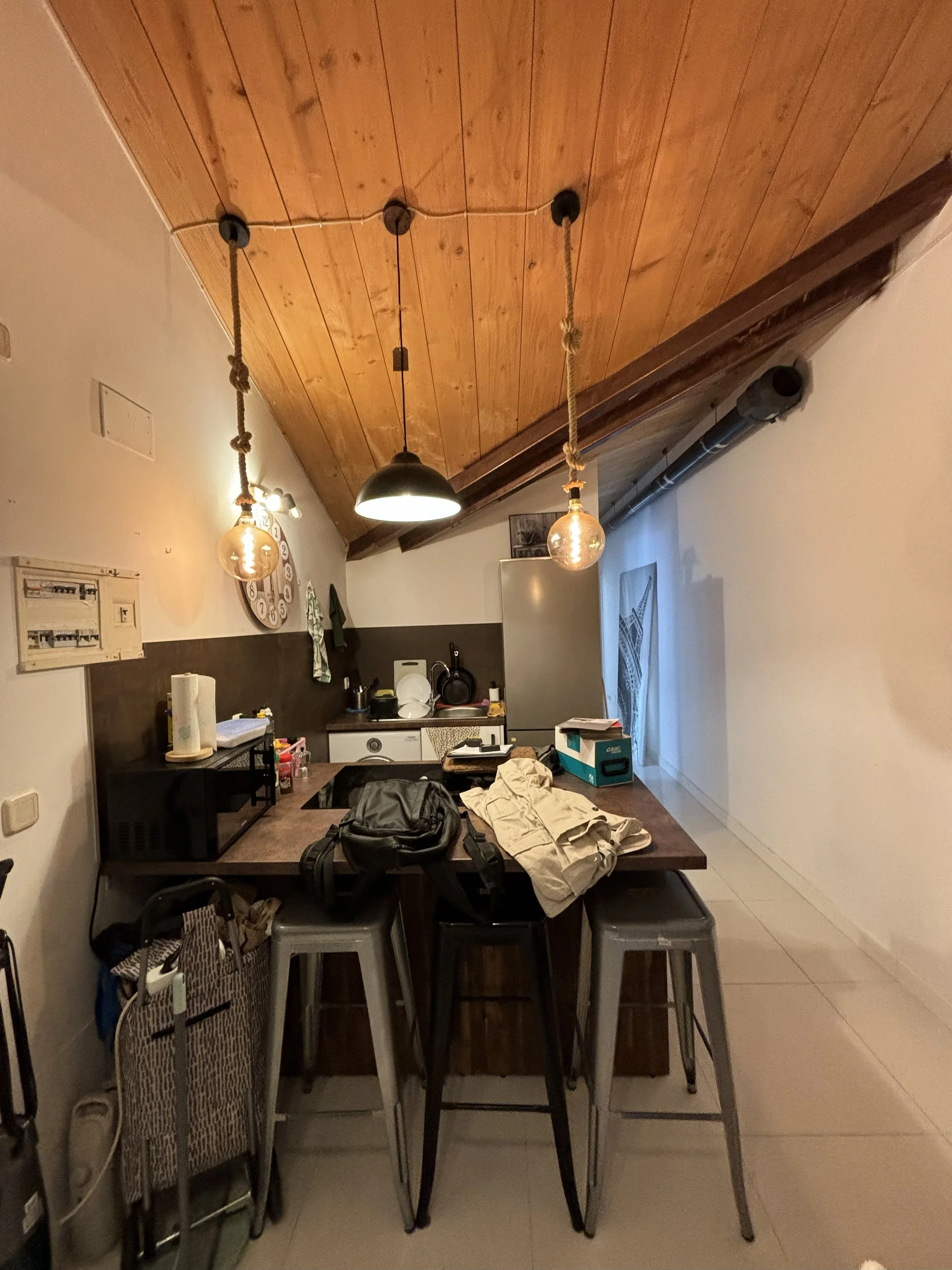 Cocina con barra y bancos de metal, techo de madera inclinada, utensilios y electrodomésticos en la encimera, lámparas colgantes con bombillas expuestas, paredes blancas, ventanas con cortinas, algunos objetos personales y bolsas sobre la mesa.