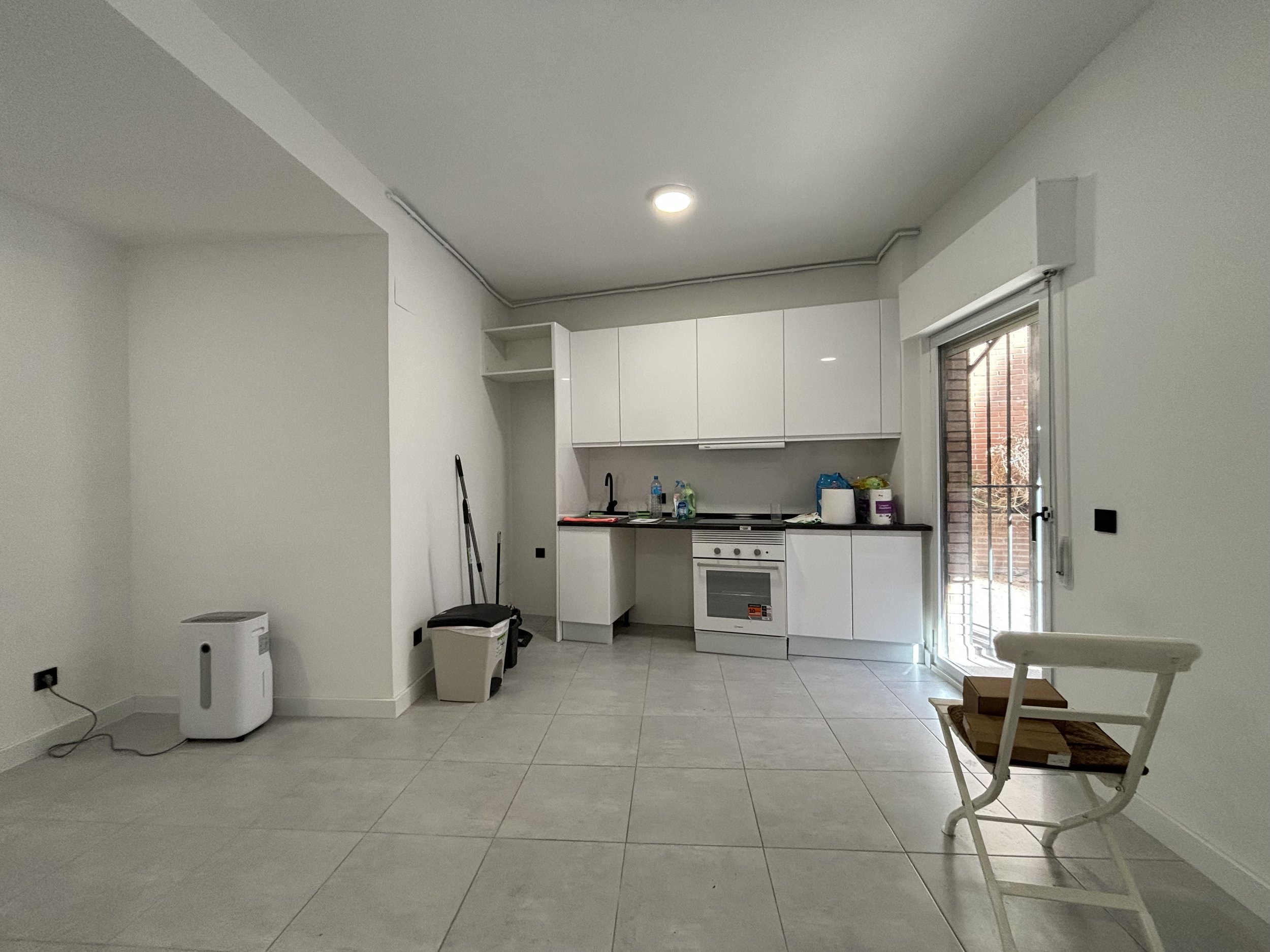 Cocina minimalista con gabinetes blancos y electrodomésticos blancos, puerta de balcón con protección de reja, piso de cerámica, y pocos objetos en la encimera.