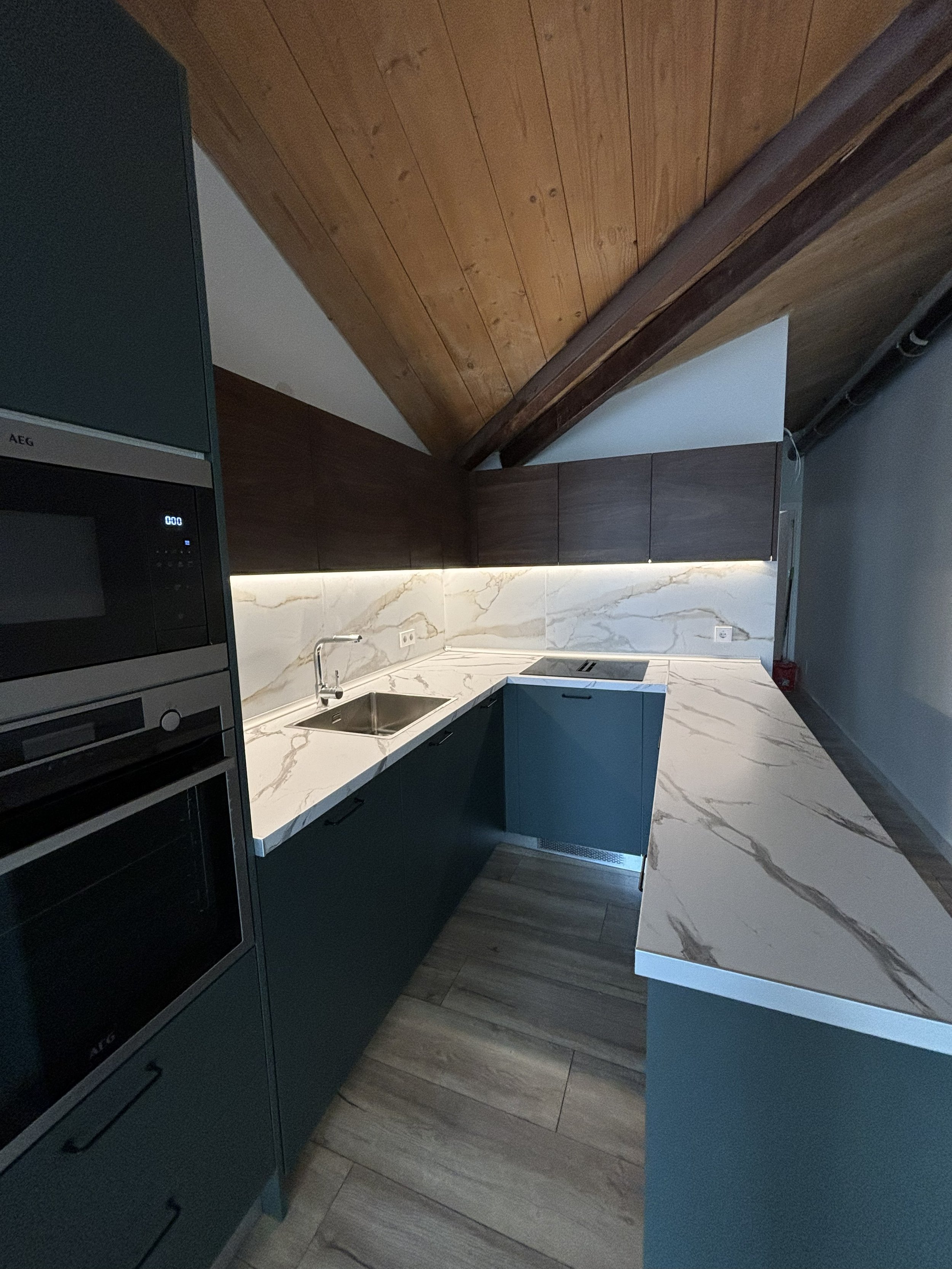 Cocina moderna con gabinetes en color verde, encimera de mármol, lavaplatos, estufa eléctrica, paredes de mármol y techo de madera en un espacio bajo el techo inclinado