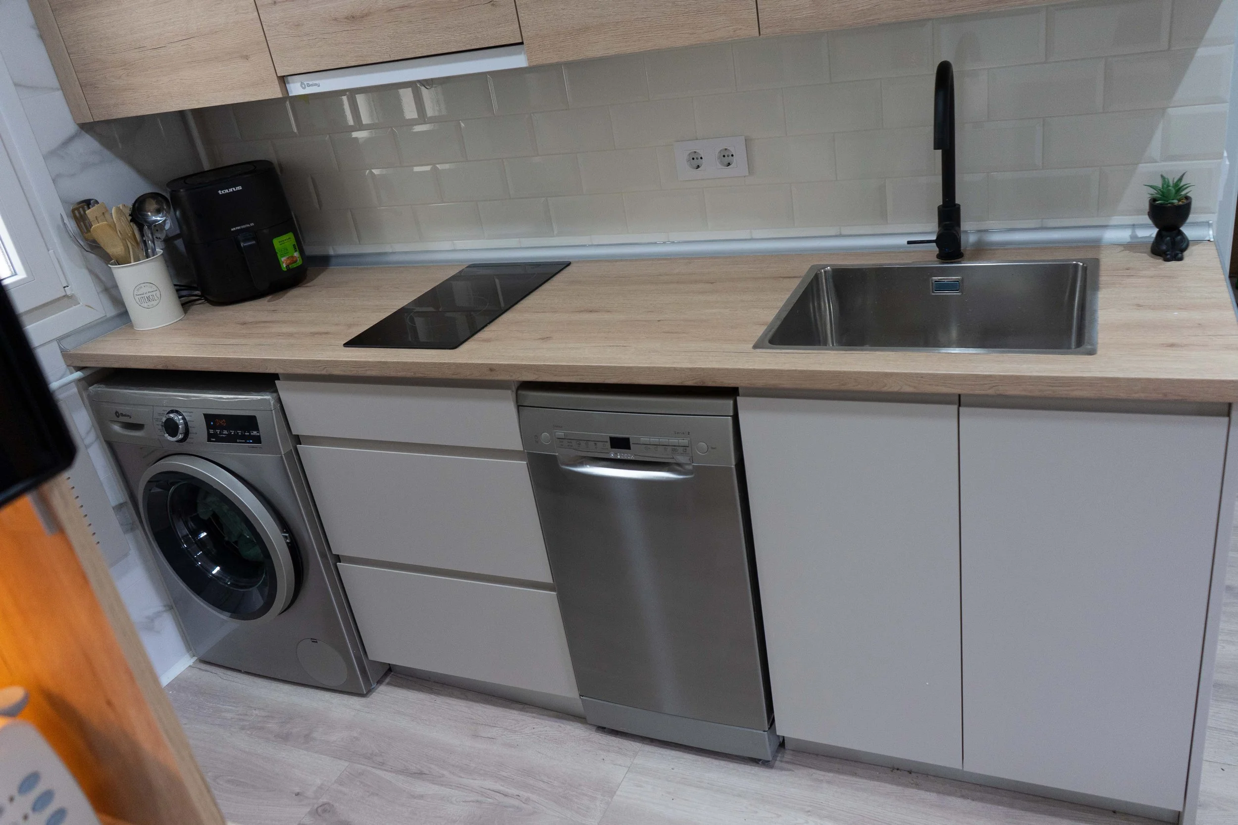 Vista de una cocina pequeña con encimera de madera, con electrodomésticos blancos y acero inoxidable, incluyendo una lavadora, un lavavajillas, y una estufa eléctrica, con un fregadero de acero inoxidable y una llave negra, y una pequeña planta en un