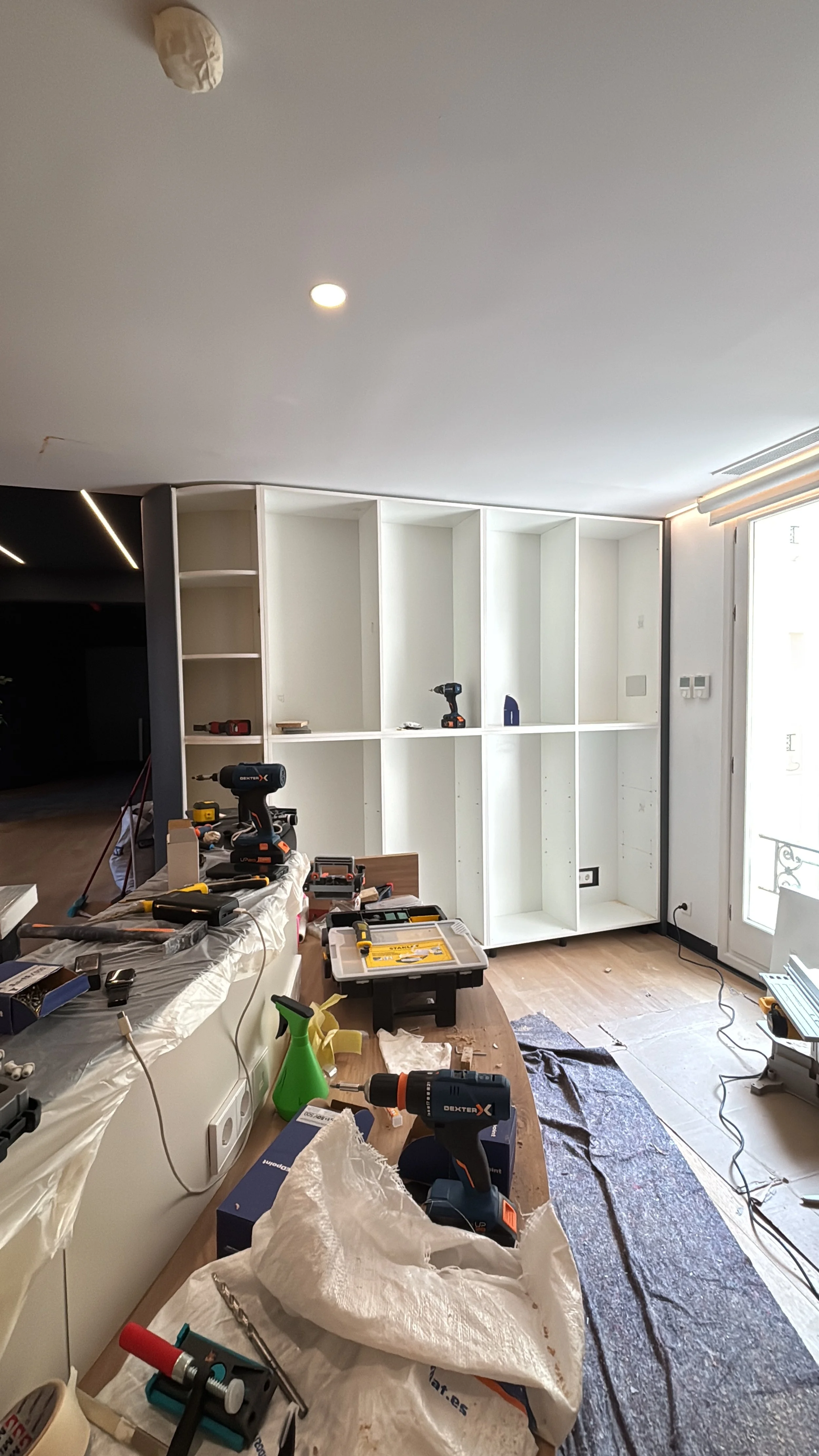 Sala en construcción con estanterías blancas, herramientas eléctricas, cables y objetos dispersos sobre una mesa de trabajo.