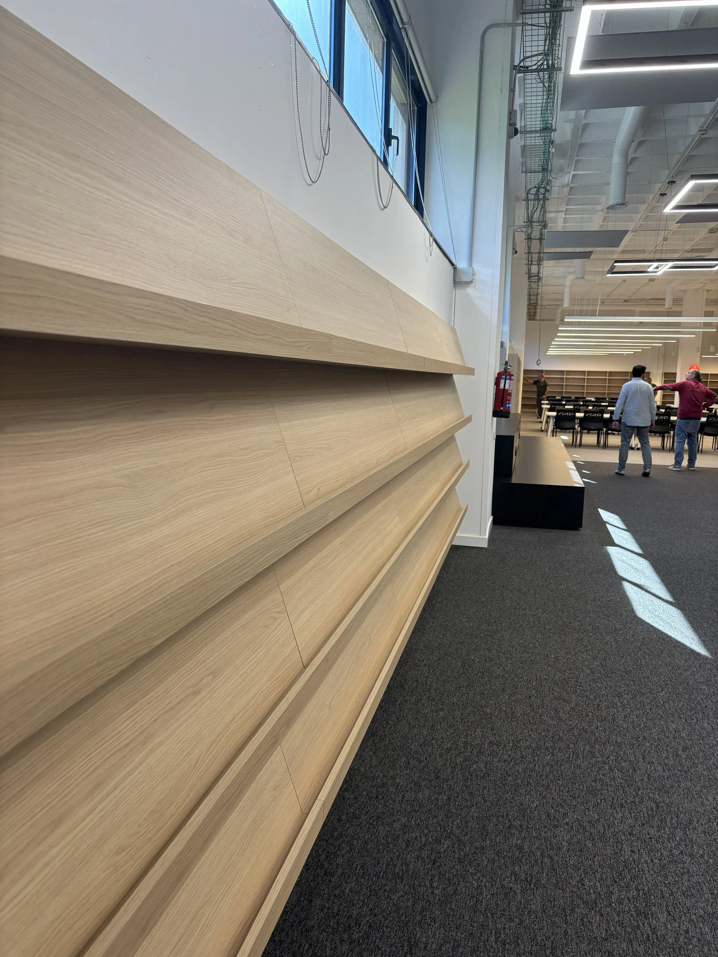 Espacio interior con paredes de madera, ventanas y personas en el fondo.