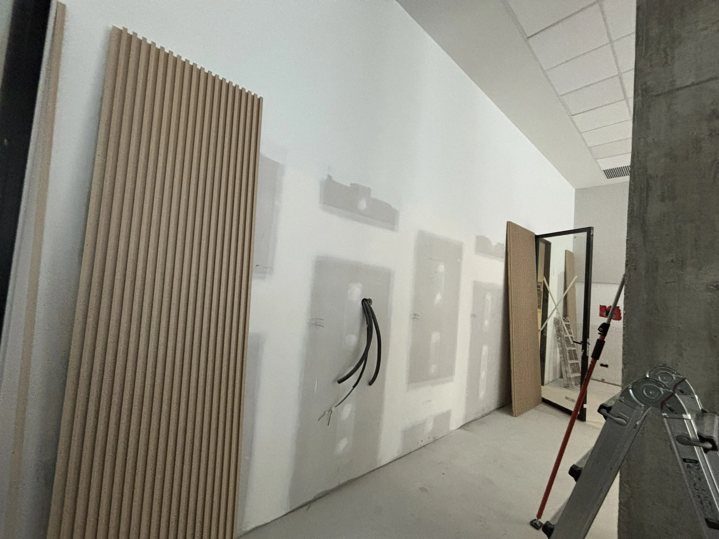 Interior en construcción con paneles de madera almacenados, cables eléctricos, un espejo de pie, escaleras y herramientas en el espacio.