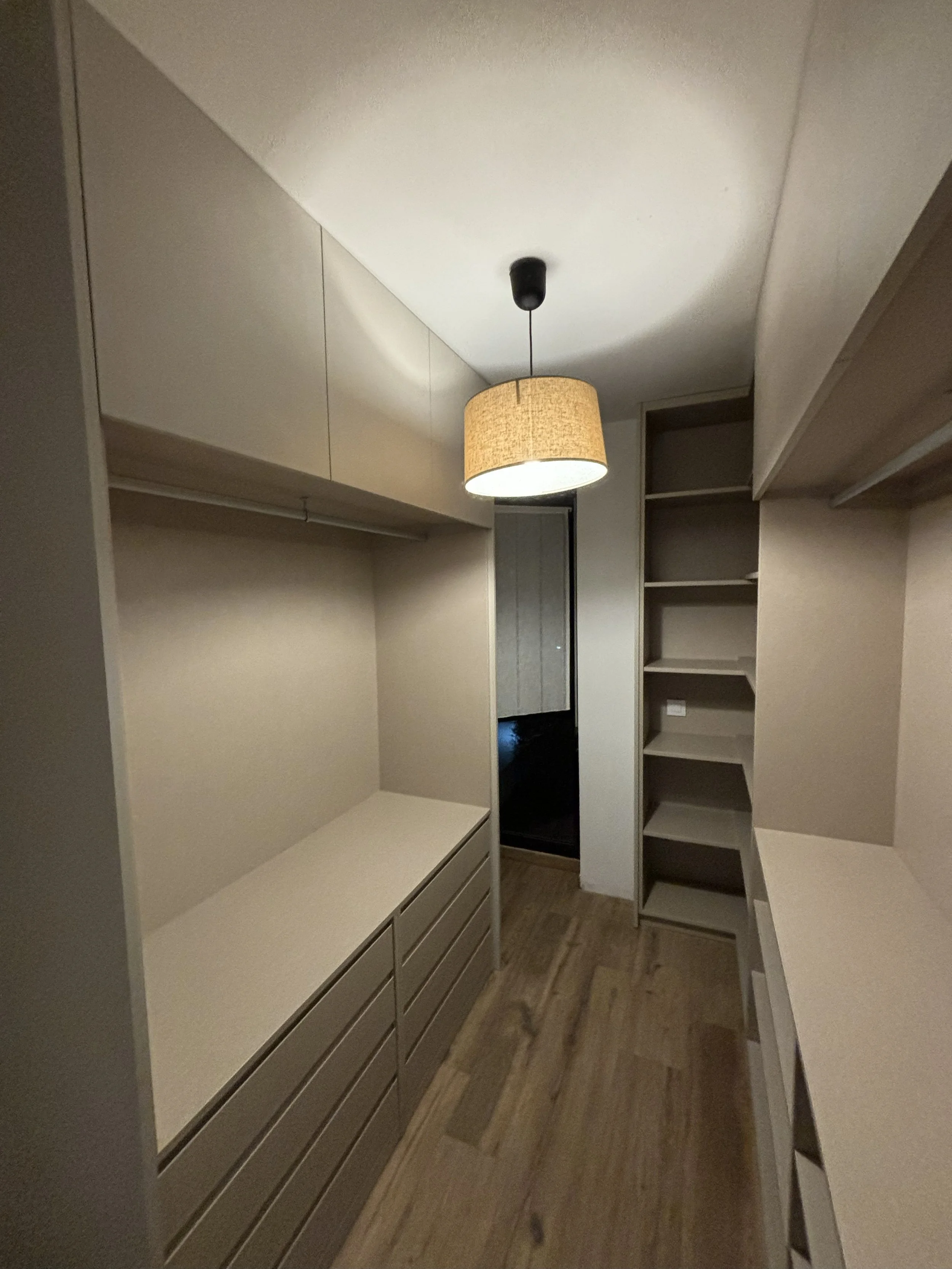 Una habitación con armario y estanterías en tonos neutros y madera, con una lámpara de techo encendida.