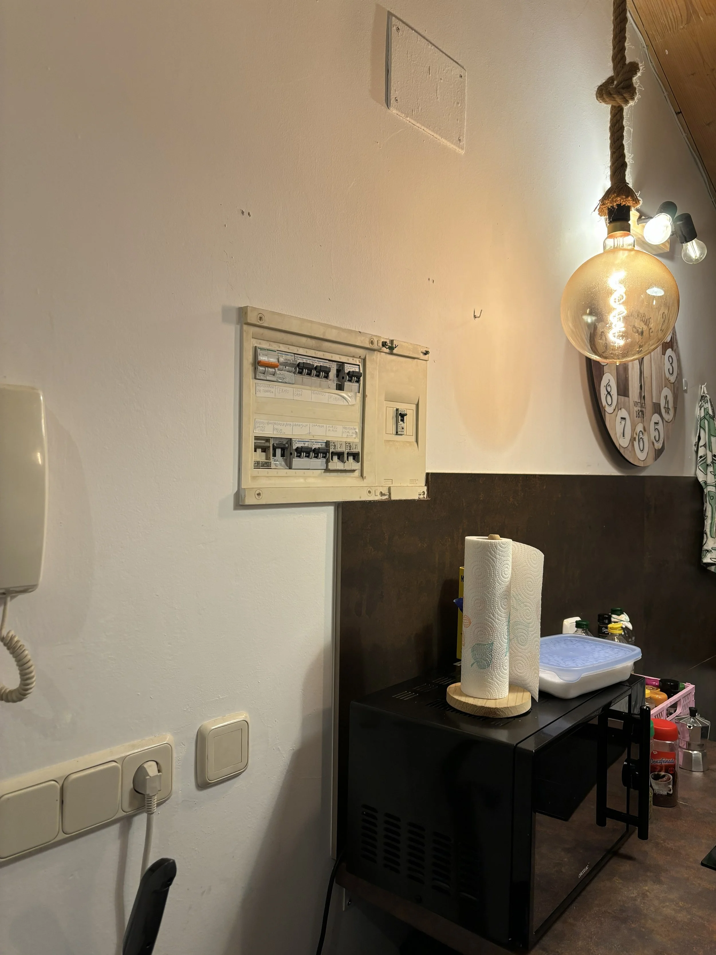 Vista de una pared con un panel de interruptores eléctricos, un reloj de pared grande, una lámpara colgante con cadena de cuerda, y una mesa con varios objetos incluyendo papel toalla, recipientes y botellas.
