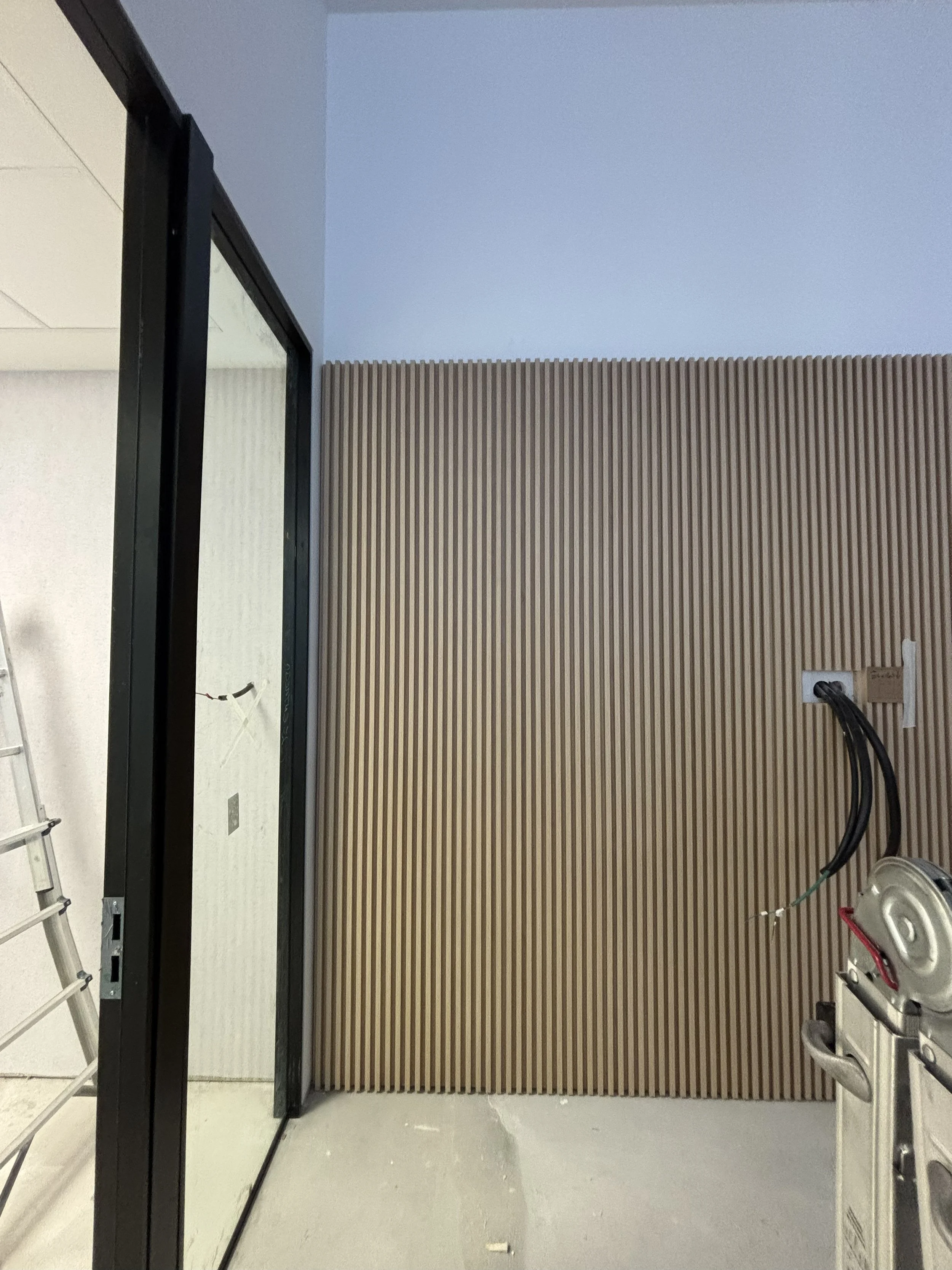 Interior de una construcción en proceso, con pared de madera de listones verticales y un enchufe con cables conectados, y una puerta de vidrio a la izquierda.