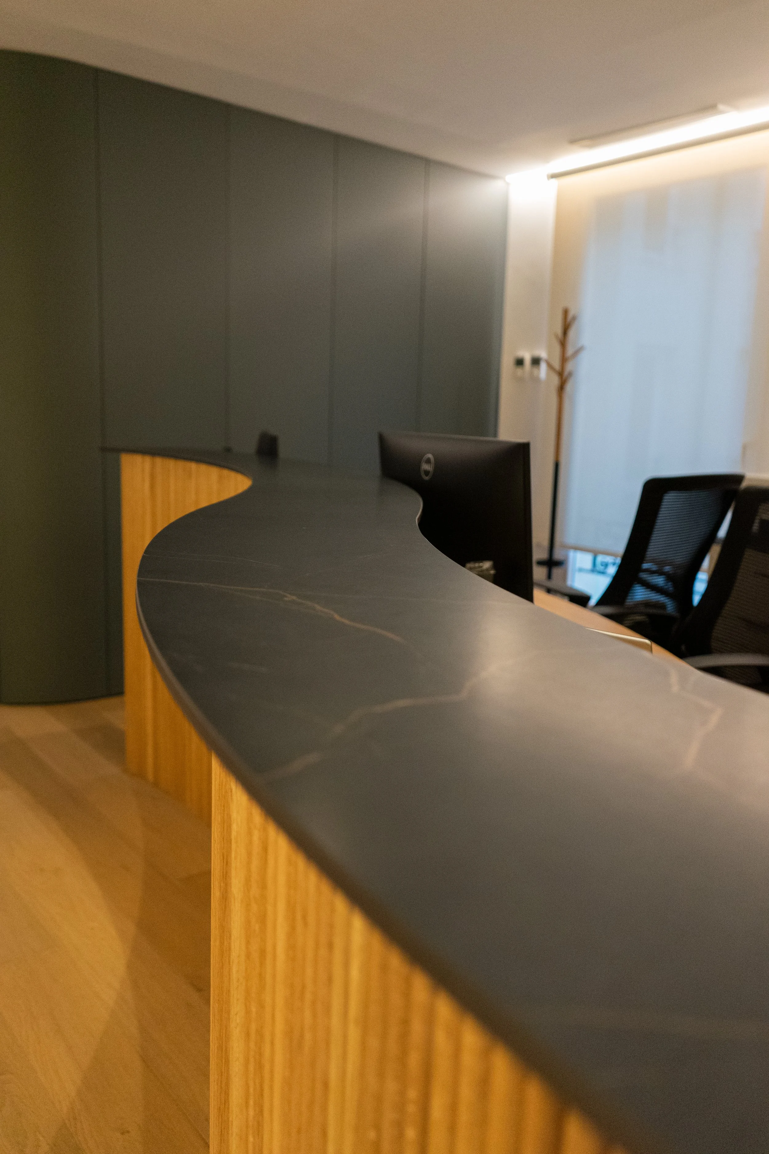 Mostrador de oficina con superficie de piedra negra y estructura de madera clara, en un ambiente con sillas y paredes de color gris.