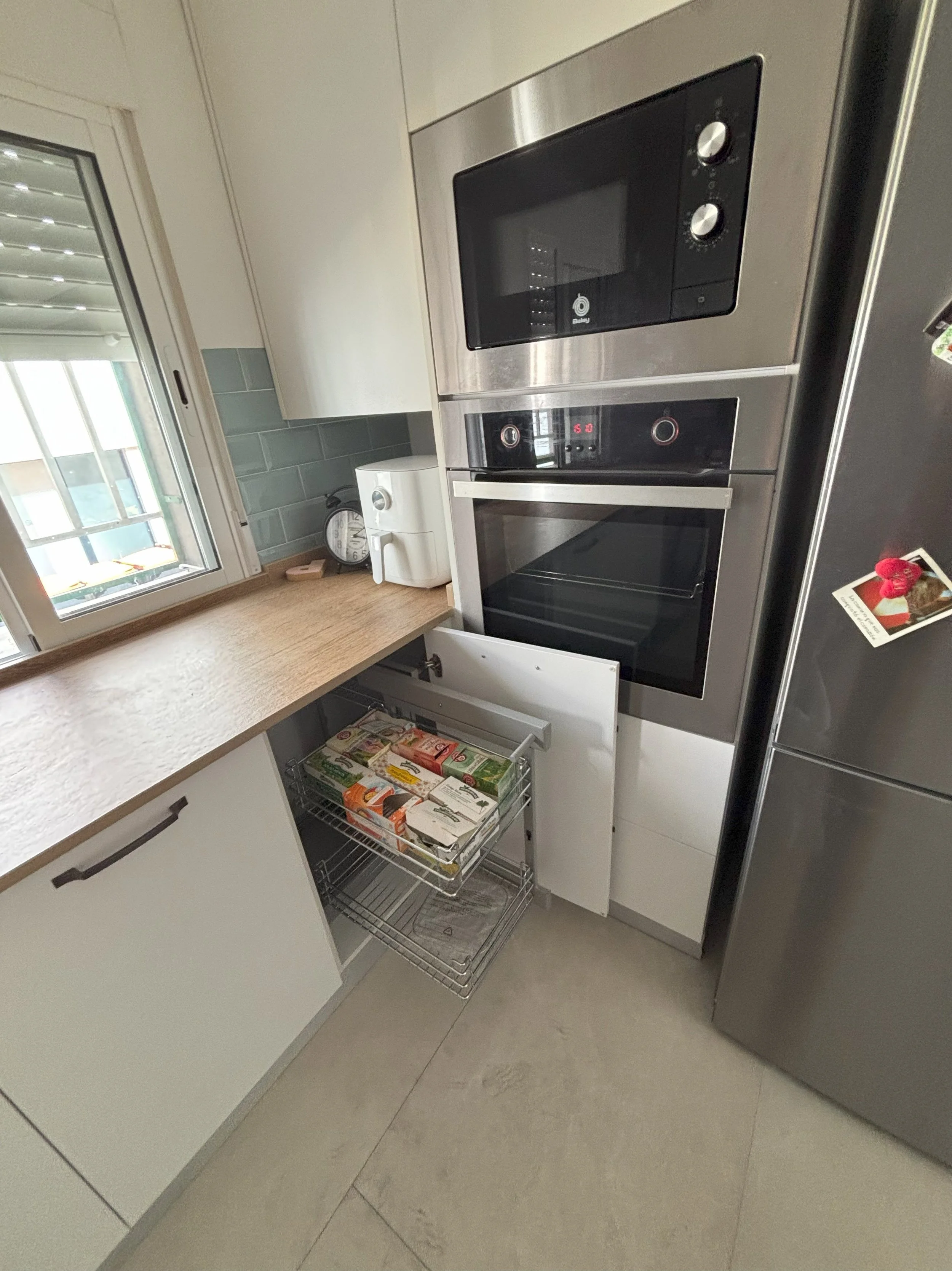 Cocina moderna con horno empotrado, microondas, refrigerator y despensa con productos en una bandeja metálica. Encimera de madera y ventana con vista exterior.