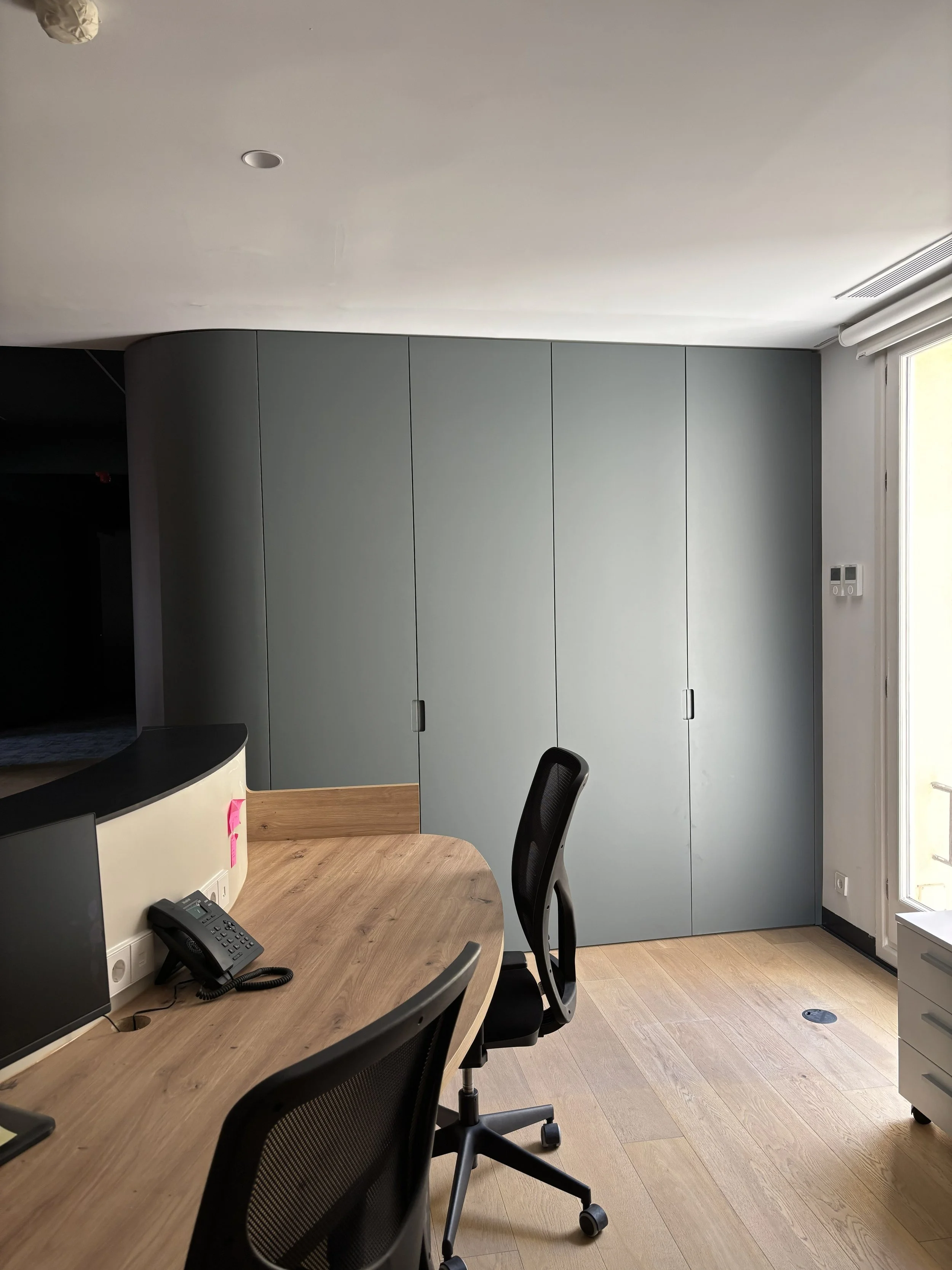 Oficina moderna con mesa de madera y sillas ergonómicas, armario gris empotrado y área de trabajo con computadora y teléfono, ventanas grandes con cortinas enrollables.