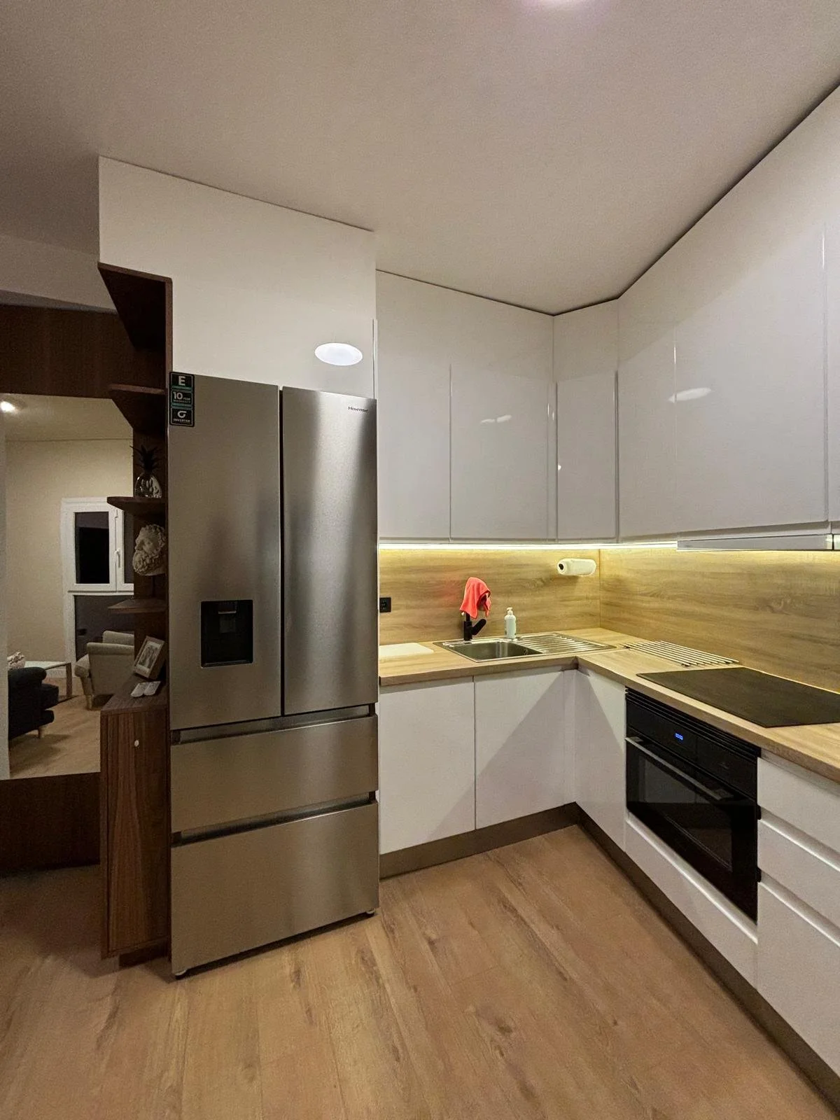 Cocina moderna con nevera de acero inoxidable, encimera de madera, gabinete blanco brillante, fregadero, estufa eléctrica y papel de cocina en la pared. Al fondo, sala con sillones y ventana.