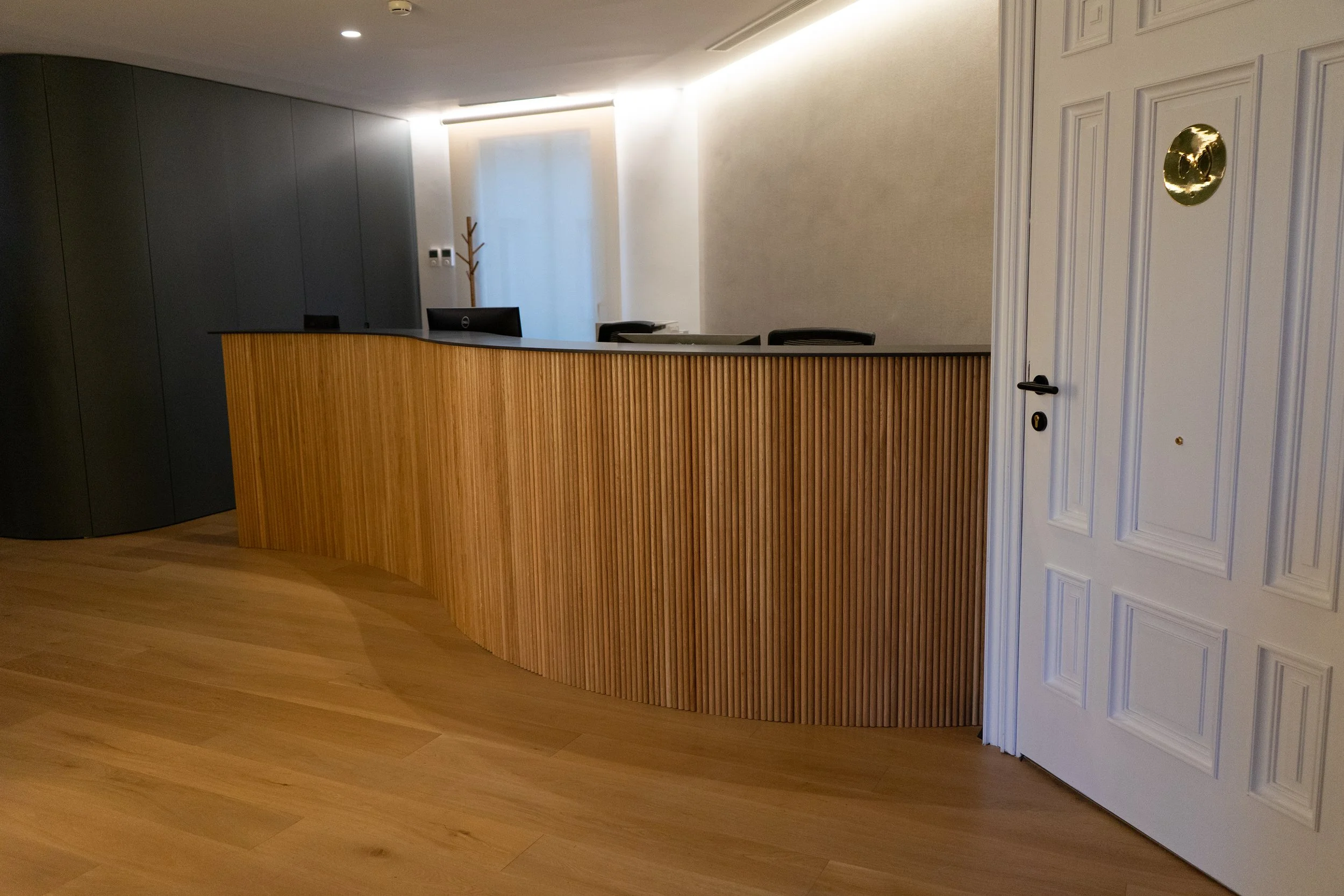 Recepción de un hotel con escritorio de madera curvada y paredes claras, con puerta blanca y piso de madera.