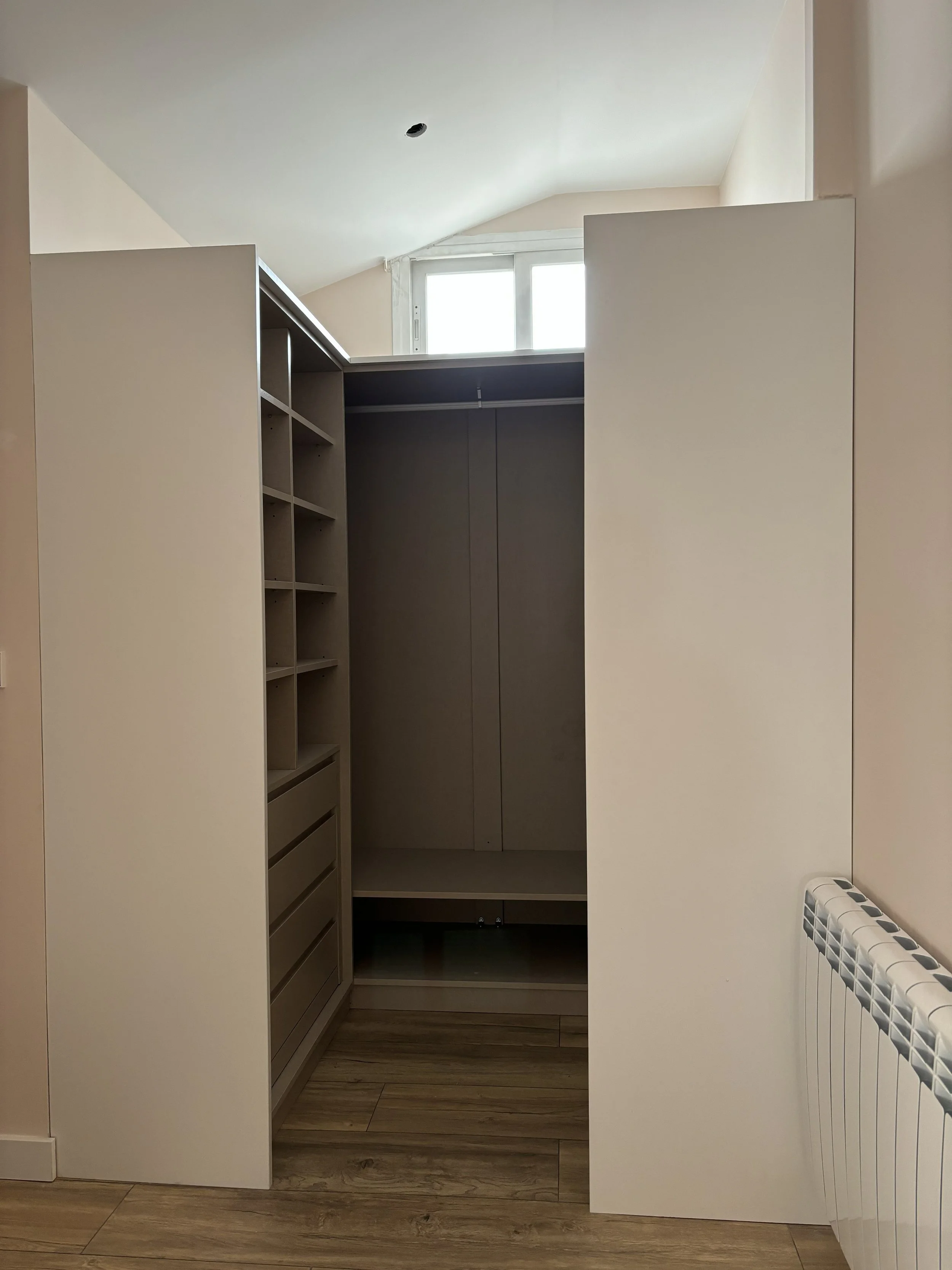 Vista de armario de madera en una habitación con suelo de madera y radiador, con ventana en el fondo y techo inclinado.