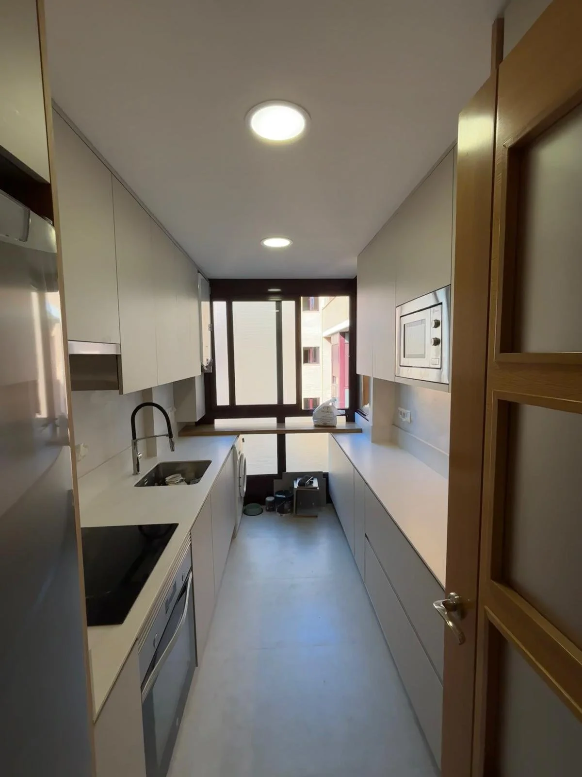 Cocina moderna y estrecha con gabinetes blancos y electrodomésticos integrados, ventana al fondo y piso de color claro.