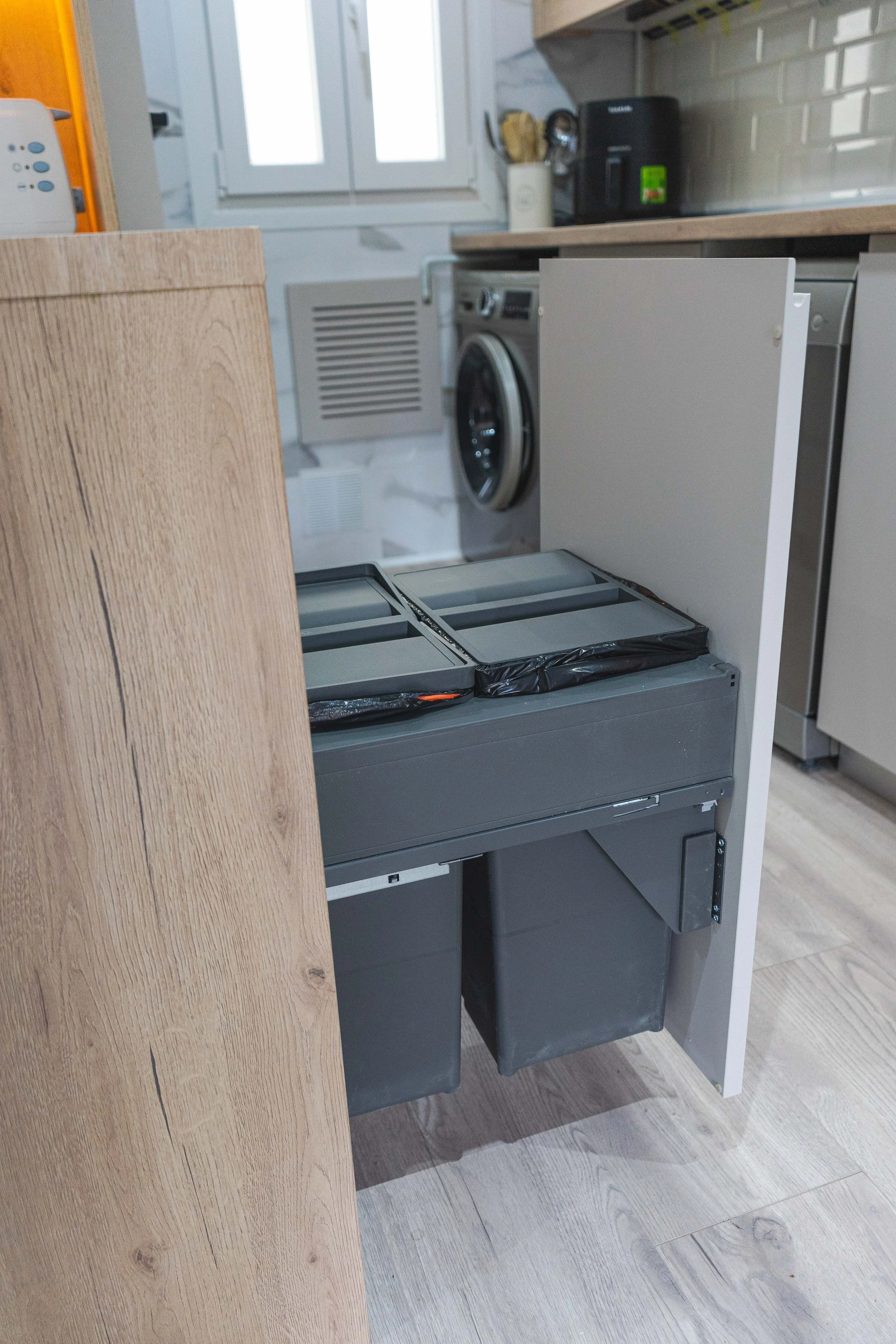 Un fregadero empotrado en una cocina moderna con gabinete de madera, con la puerta del mueble abierta mostrando un contenedor de basura y un lavabo.