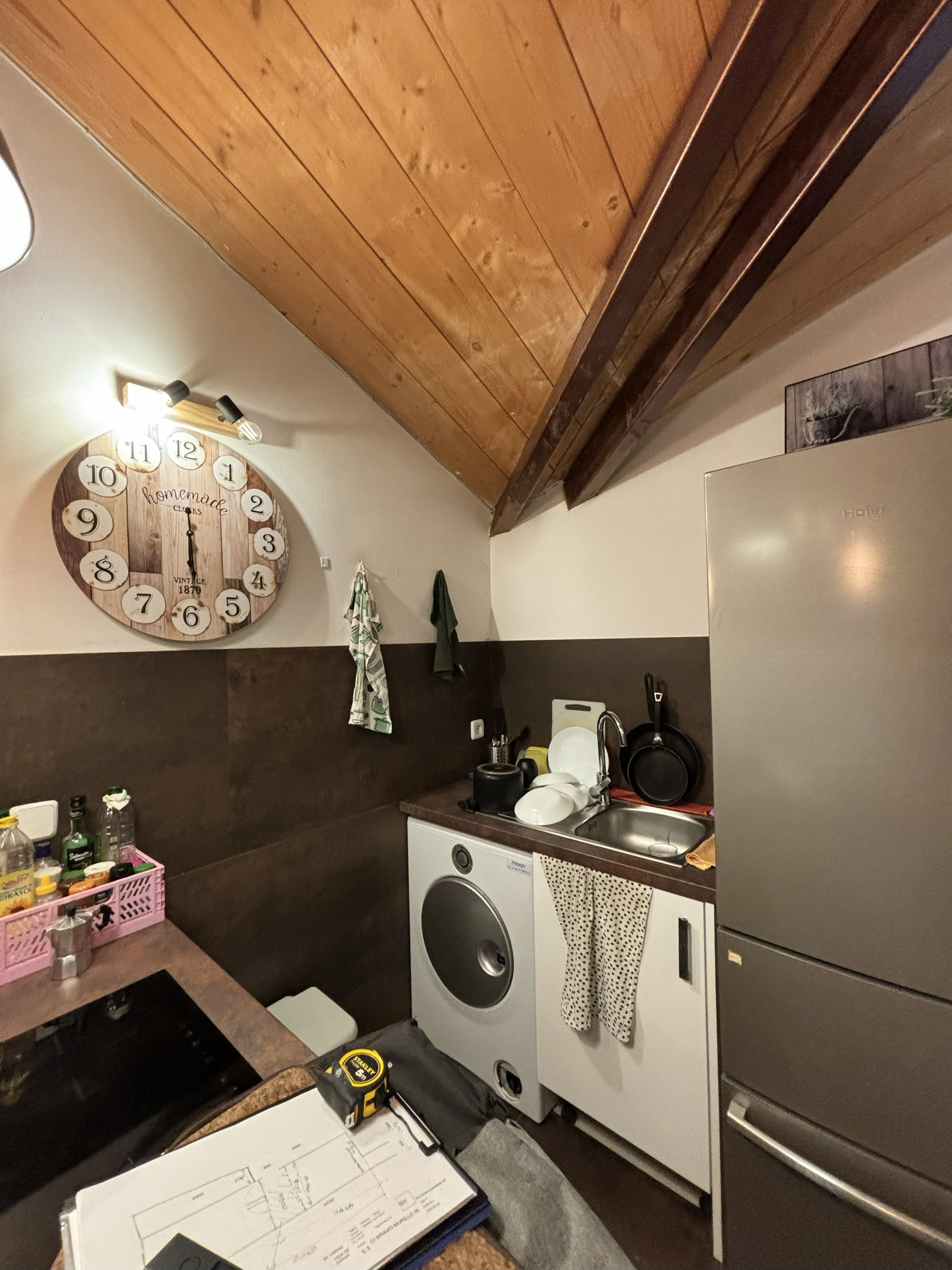 Cocina en un ático con paredes de madera y techos inclinados. Incluye una nevera grande, un fregadero, un lavavajillas, y una mesa con instrumentos y papeles.