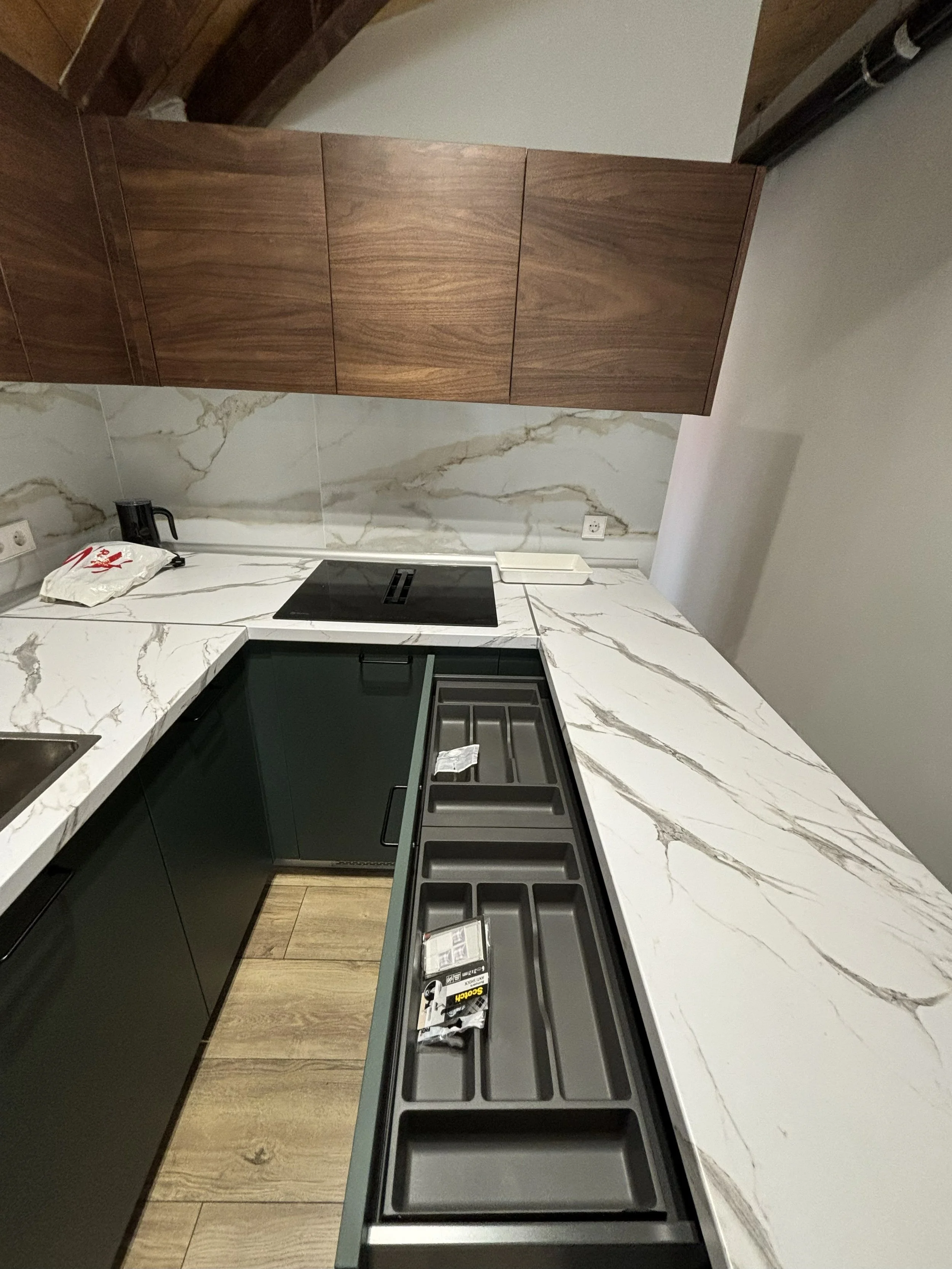 Cocina moderna con encimera de mármol blanco con vetas grises, gabinetes oscuros, una estufa eléctrica, y armarios de madera sobre la encimera. Hay objetos pequeños y un trapo en la encimera.