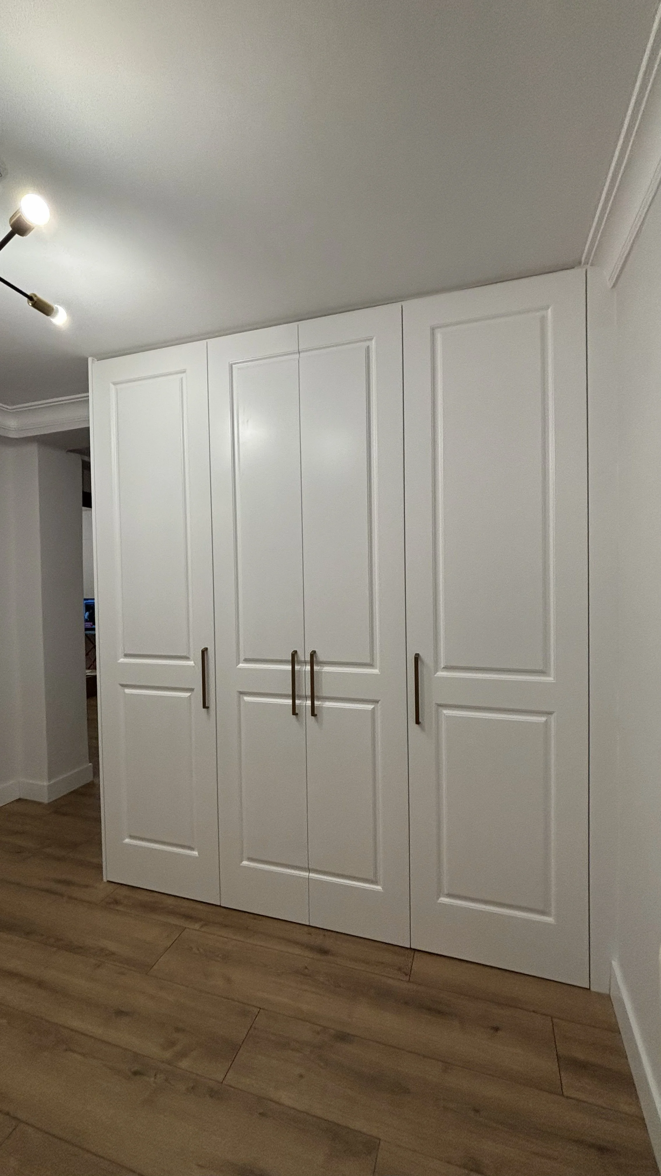 Armario blanco de closet con puertas de panel y tiradores metálicos de color dorado, en una habitación con suelo de madera y paredes blancas.