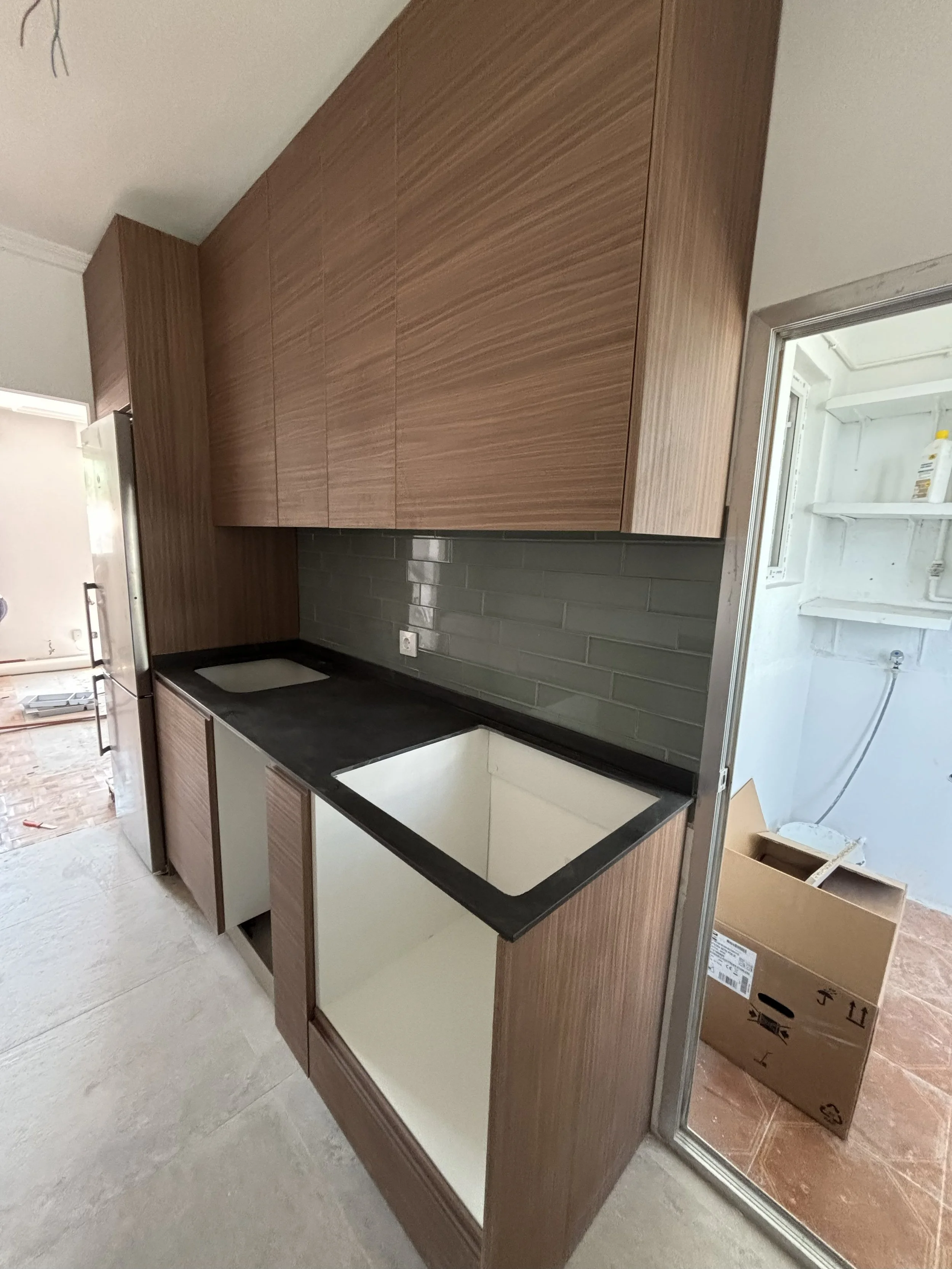 Cocina en construcción con gabinetes de madera y una encimera negra, fregadero sin instalar, y fondo de paredes con azulejos en color gris oscuro.