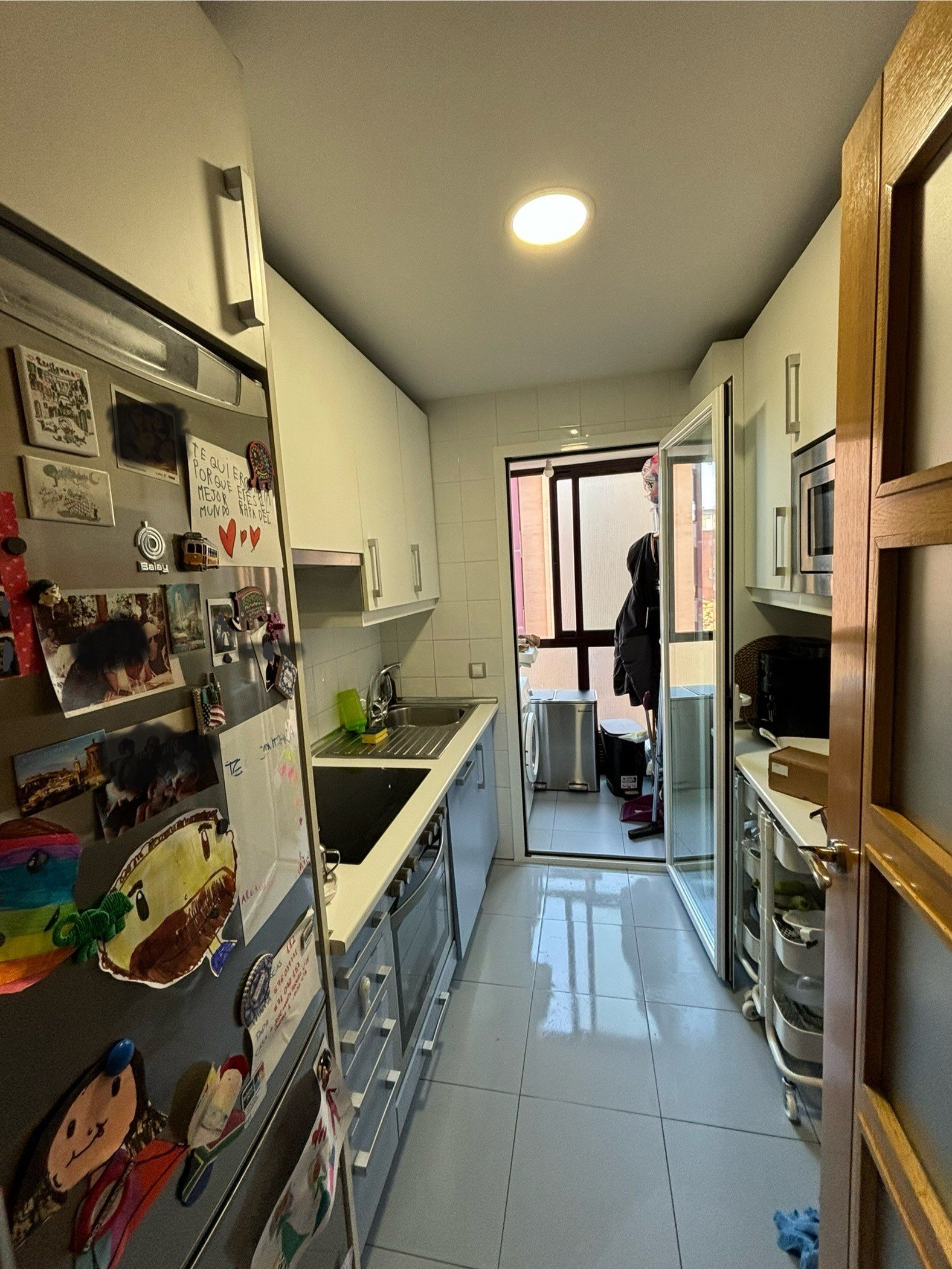 Cocina moderna con gabinetes blancos, refrigerador decorado con fotos y notas, cocina eléctrica, lavabo, mini balcón al fondo con ventana y persona mirando hacia afuera.