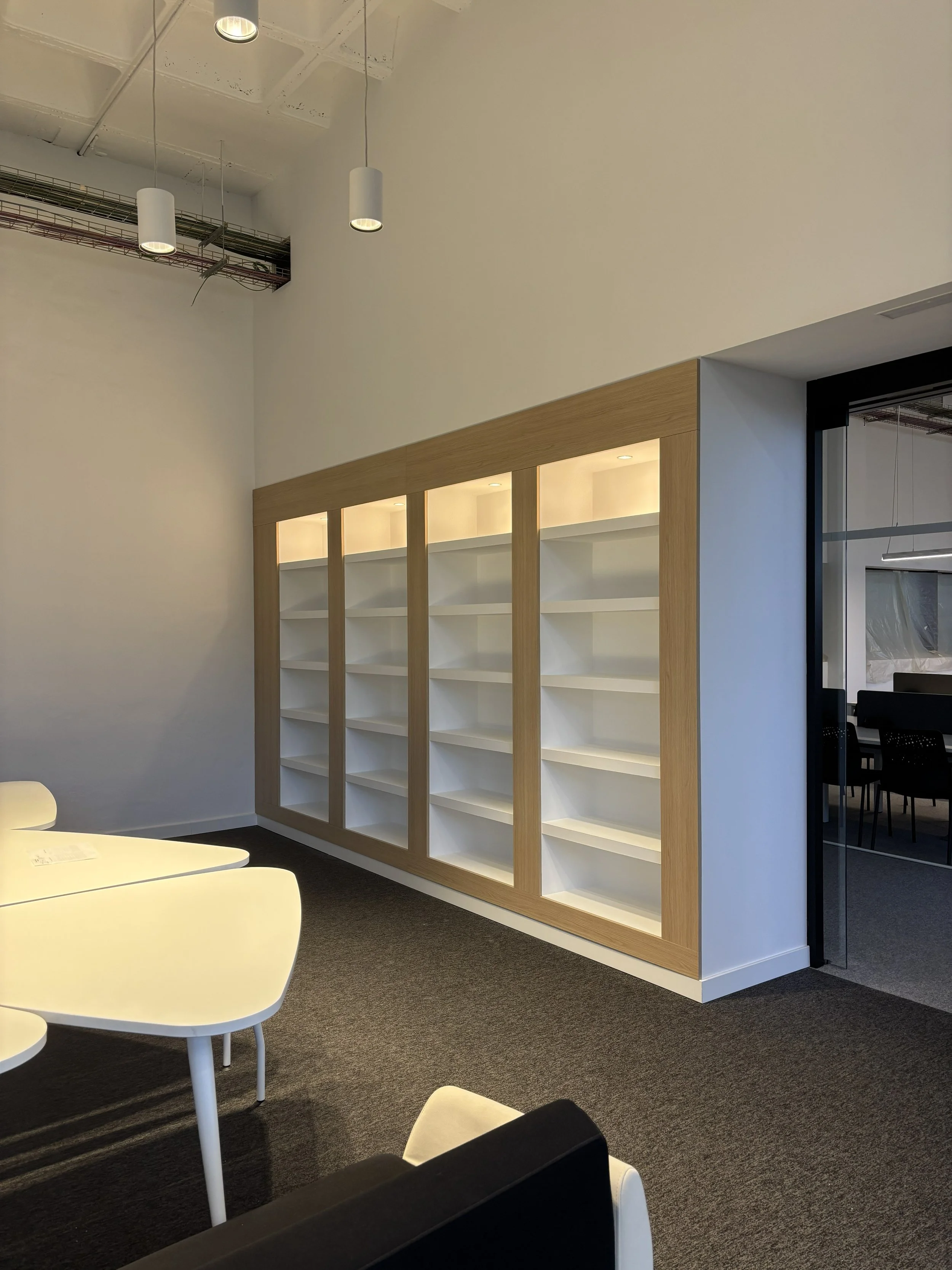 Una sala moderna con una pared de estanterías blancas y marco de madera, una mesa blanca y sillas negras y blancas. Ventana de acrílico con puerta de cristal a la derecha.