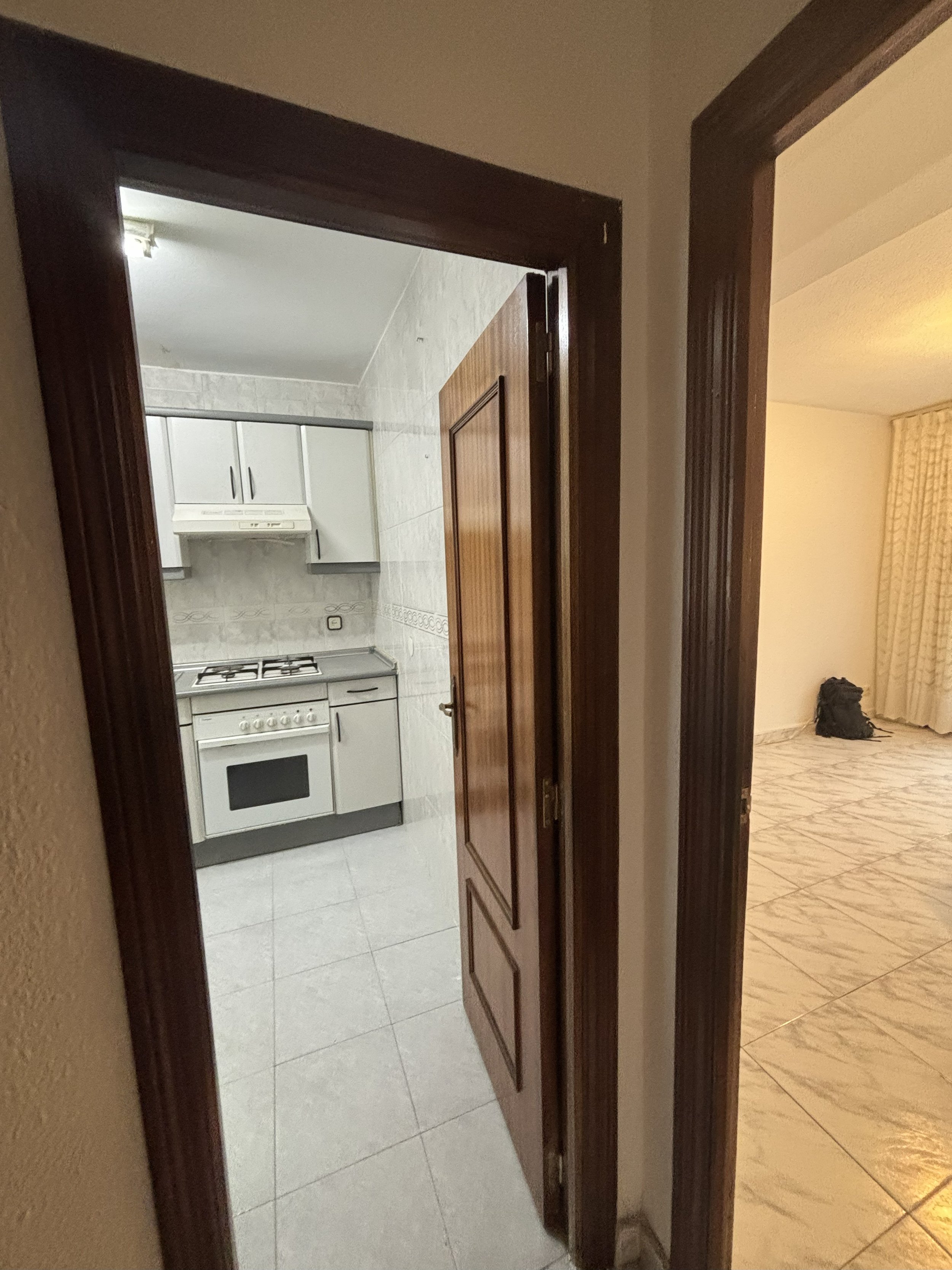 Vista de una cocina con gabinetes blancos y estufa, vista desde una entrada, con un pasillo a la derecha y una sala de estar con cortinas en el fondo.