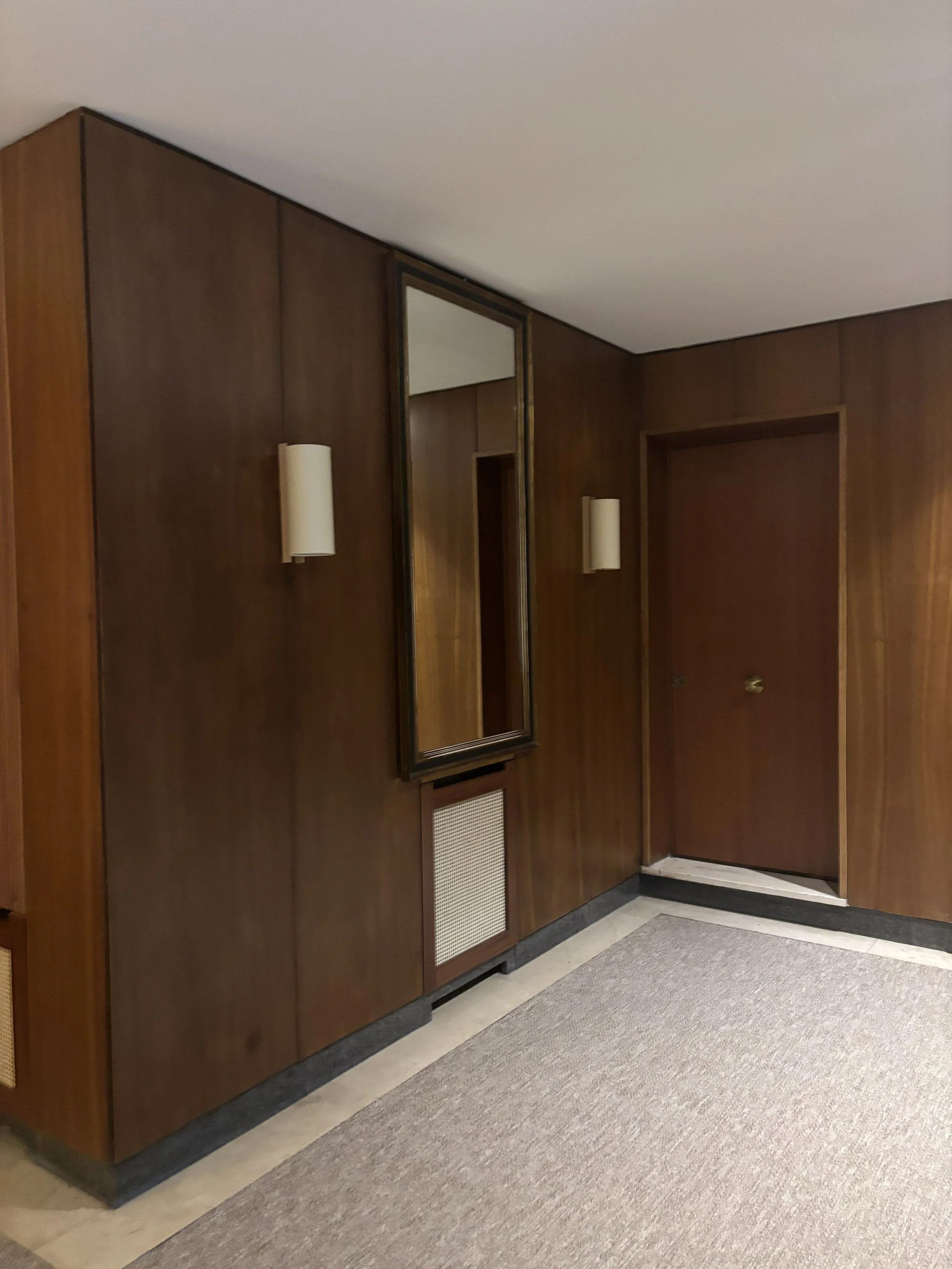 Pared de madera en un interior, con un espejo rectangular, dos apliques de luz, y una puerta cerrada.