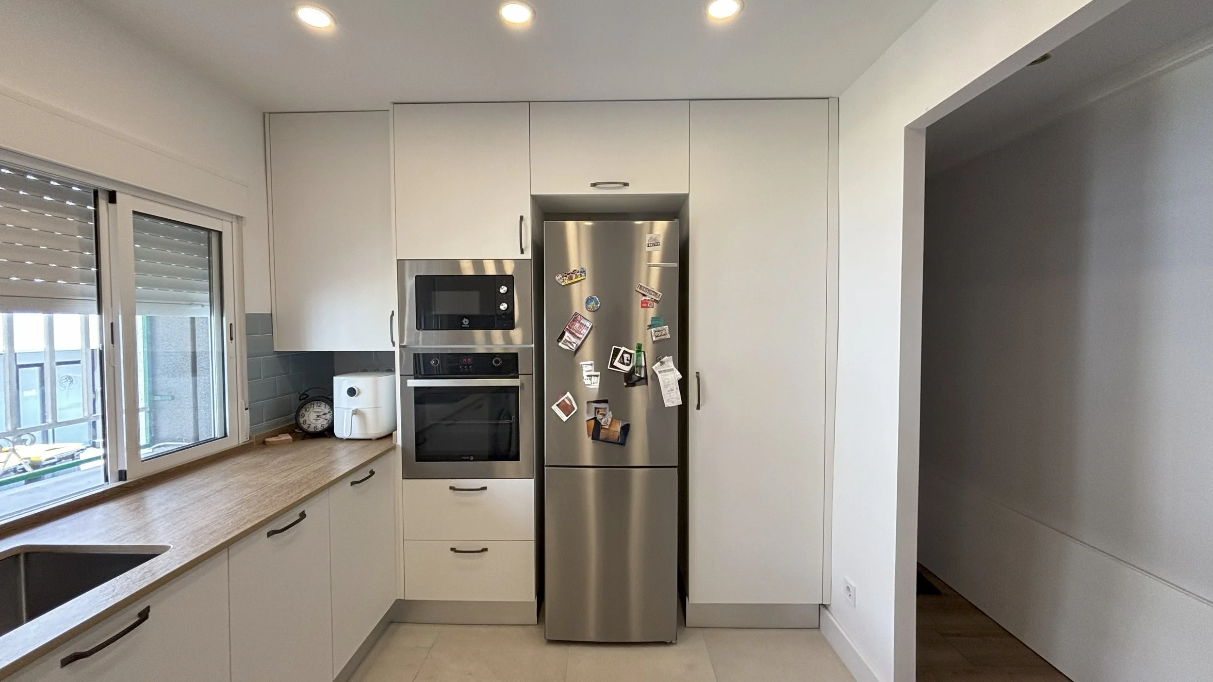 Cocina moderna con ventanas, armarios blancos, electrodomésticos de acero inoxidable y un refrigerador cubierto con imanes y notas.