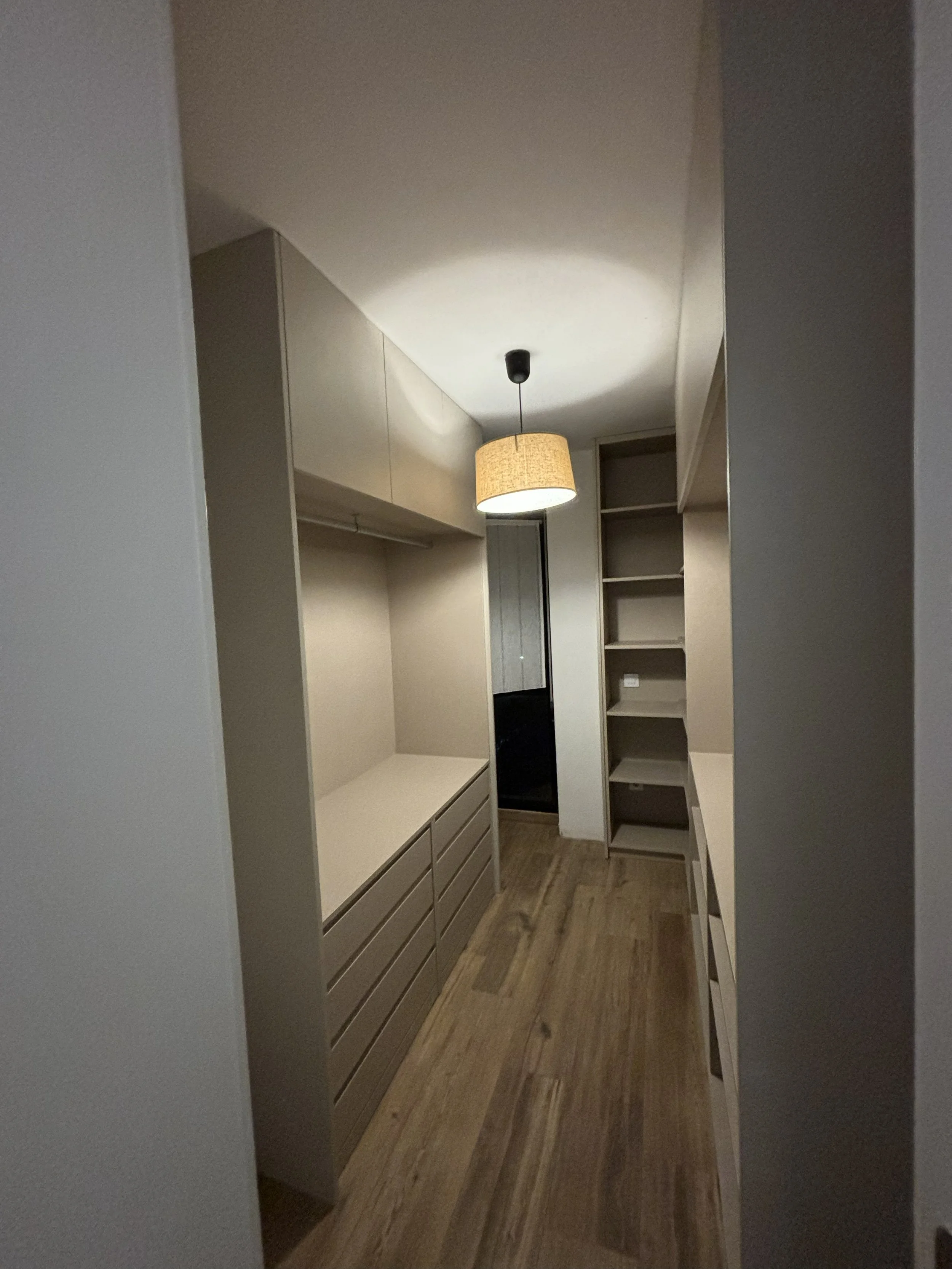 Es un vestidor con armarios blancos, un estante, y un espacio para colgar ropa. El piso es de madera y hay una lámpara colgante sobre el vestidor.