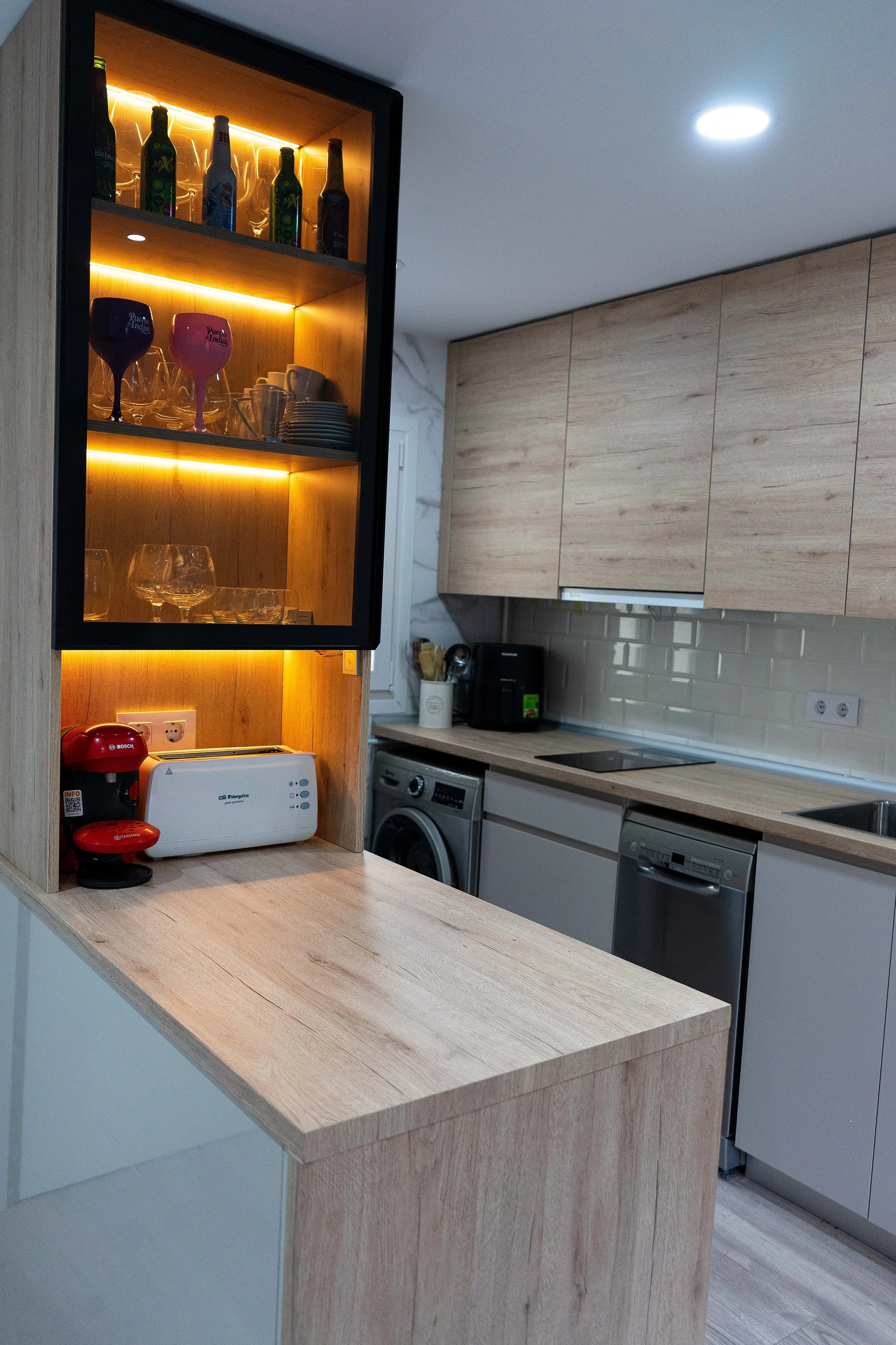 Cocina moderna con electrodomésticos de acero, gabinetes de madera clara, estantes con cristalería y bebidas, y accesorios de cocina en la encimera.