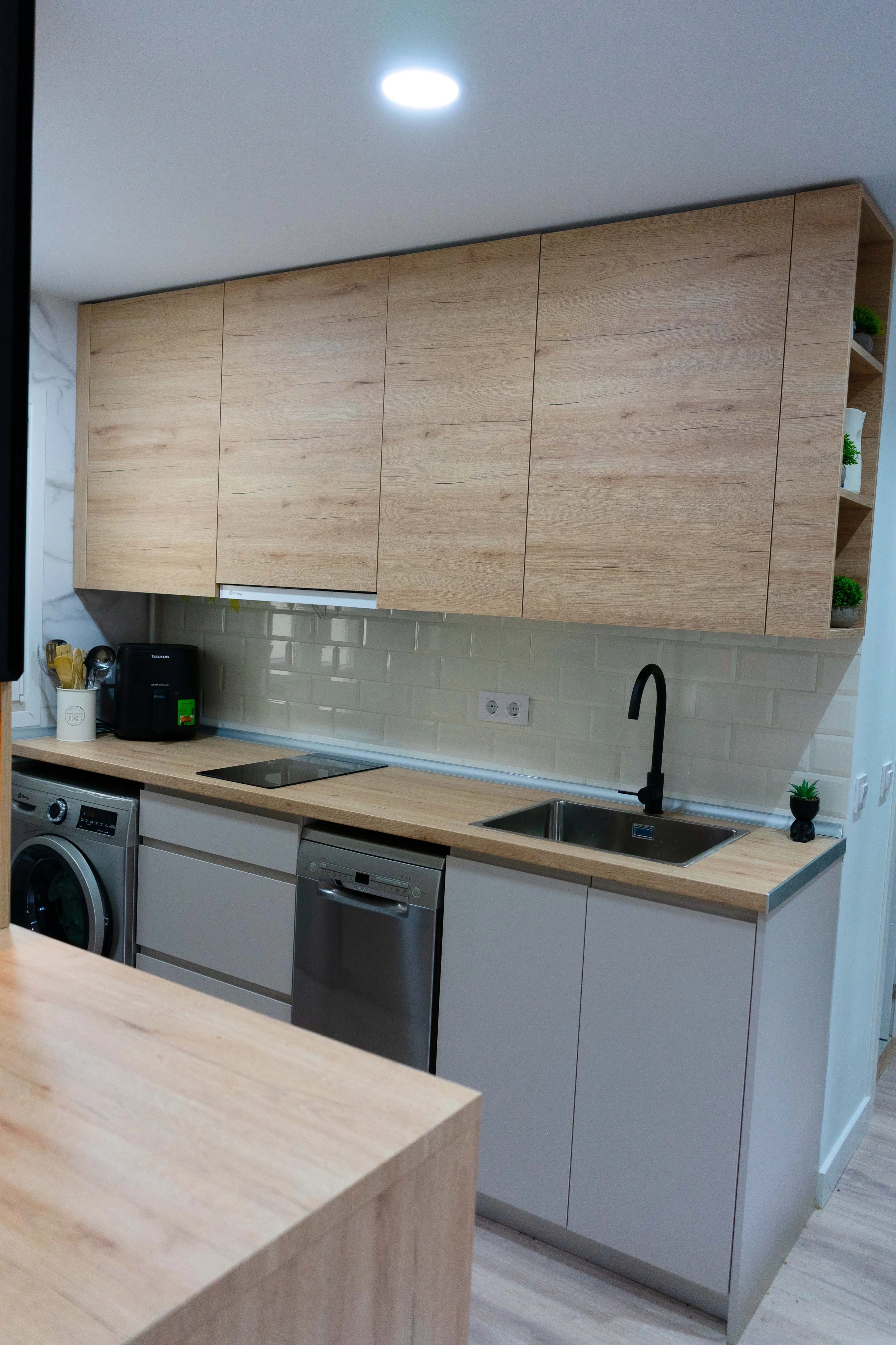 Cocina moderna con gabinetes de madera clara, encimera de madera, fregadero con grifo negro, electrodomésticos de acero inoxidable, azulejos beige en la pared y pequeños accesorios decorativos con plantas artificiales.