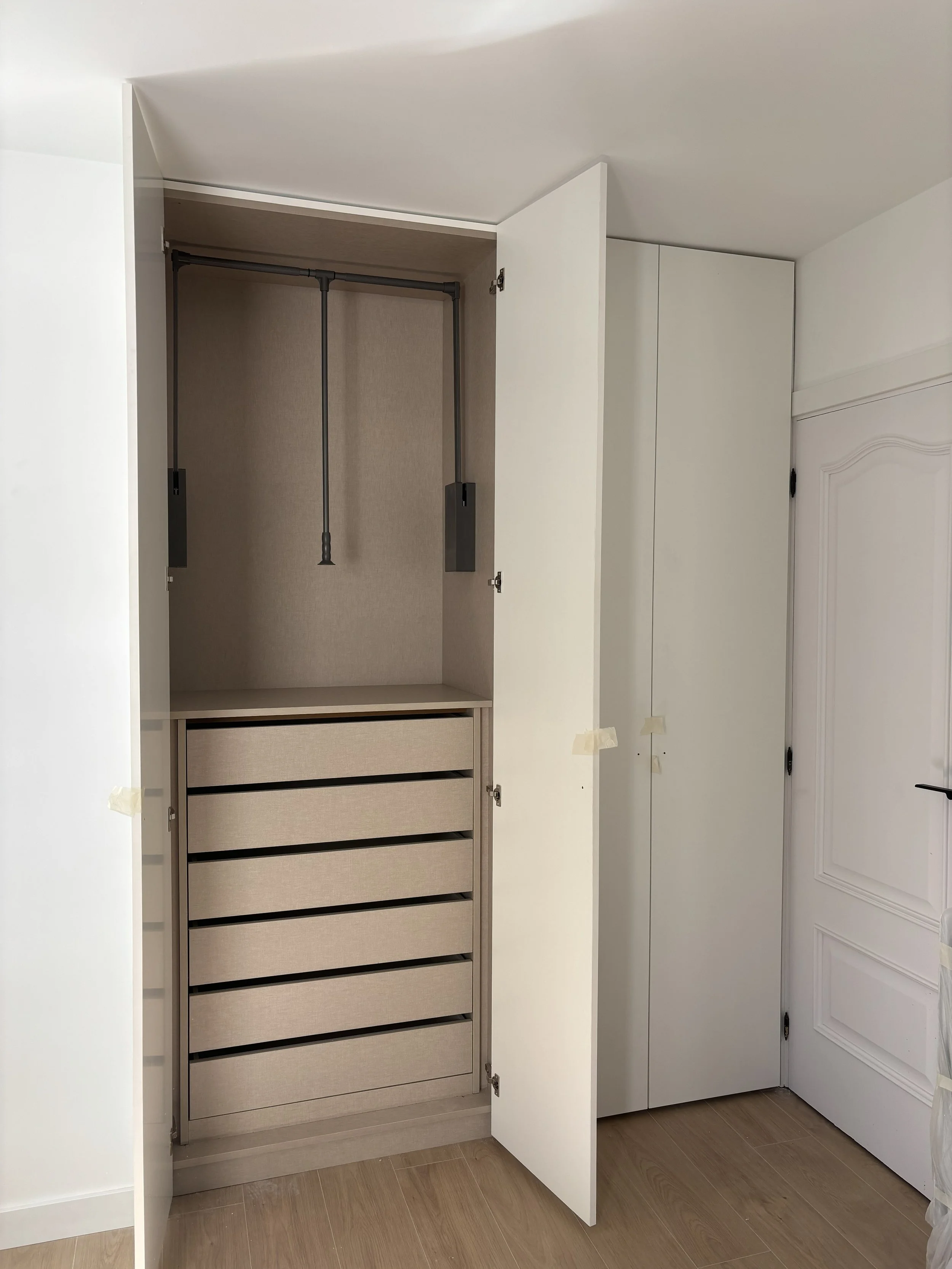 Armario empotrado con puertas abiertas, vacío, con estructura para colgar ropa y gavetas de almacenamiento al fondo, en una habitación con piso de madera y puerta blanca decorativa.