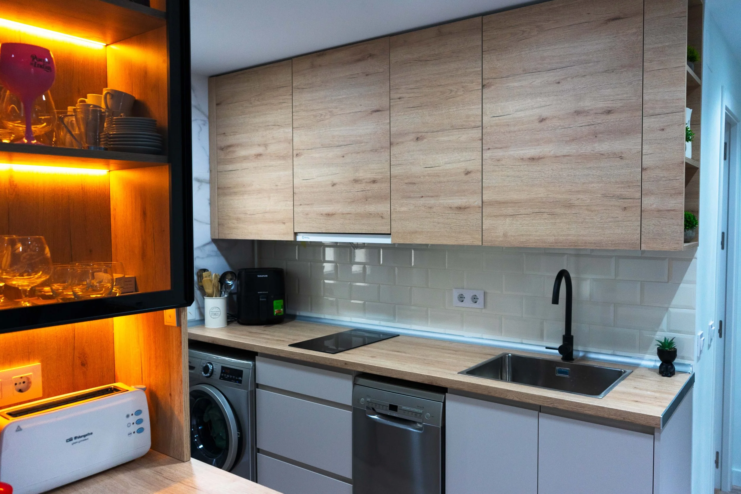 Cocina moderna con gabinetes de madera clara, electrodomésticos de acero, incluyendo lavavajillas, horno de microondas y una estufa eléctrica, y decoración con pequeñas plantas.
