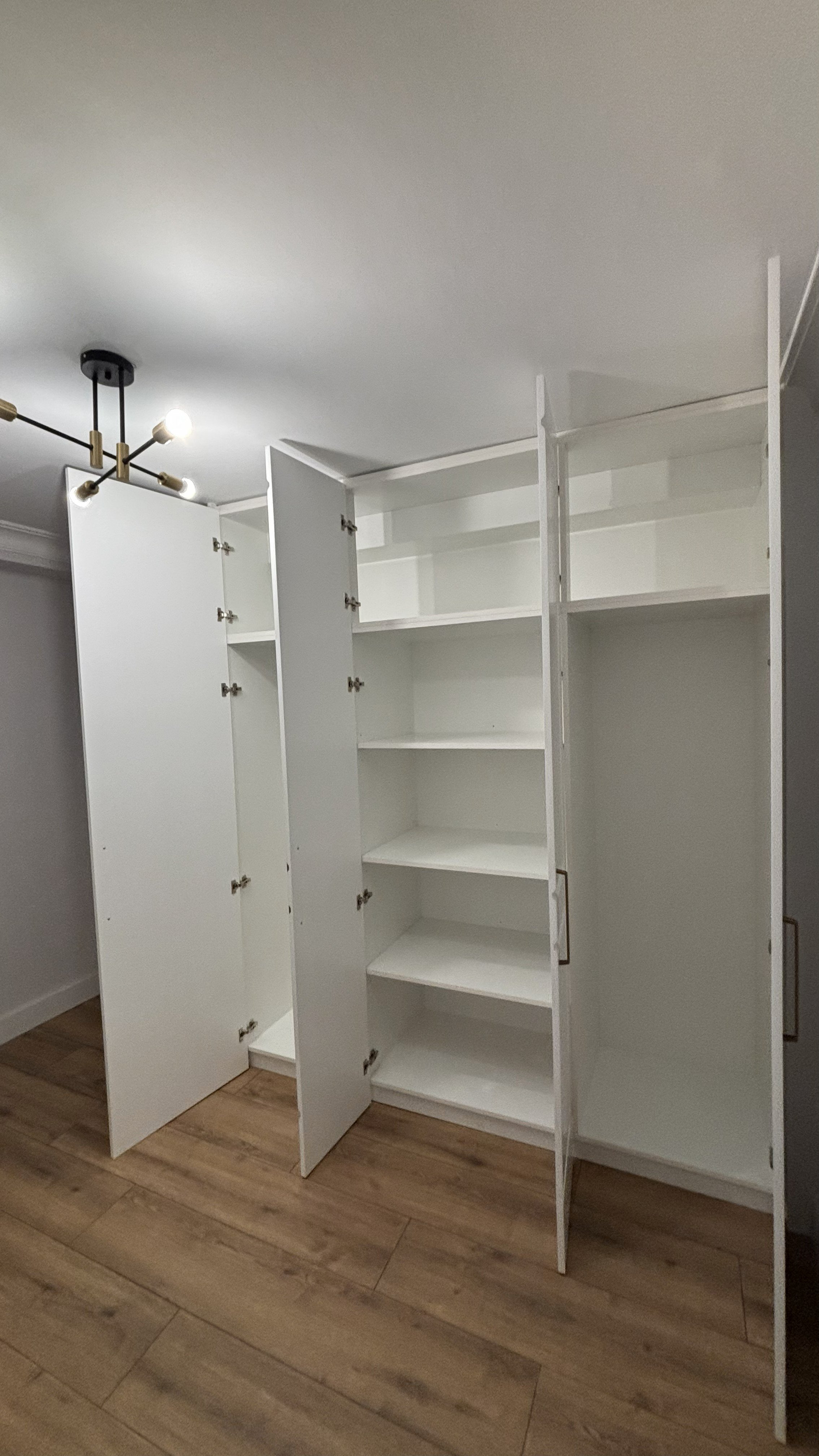 Armario blanco con puertas abiertas, mostrando estantes en el interior, en una habitación con piso de madera y luz de techo moderna.