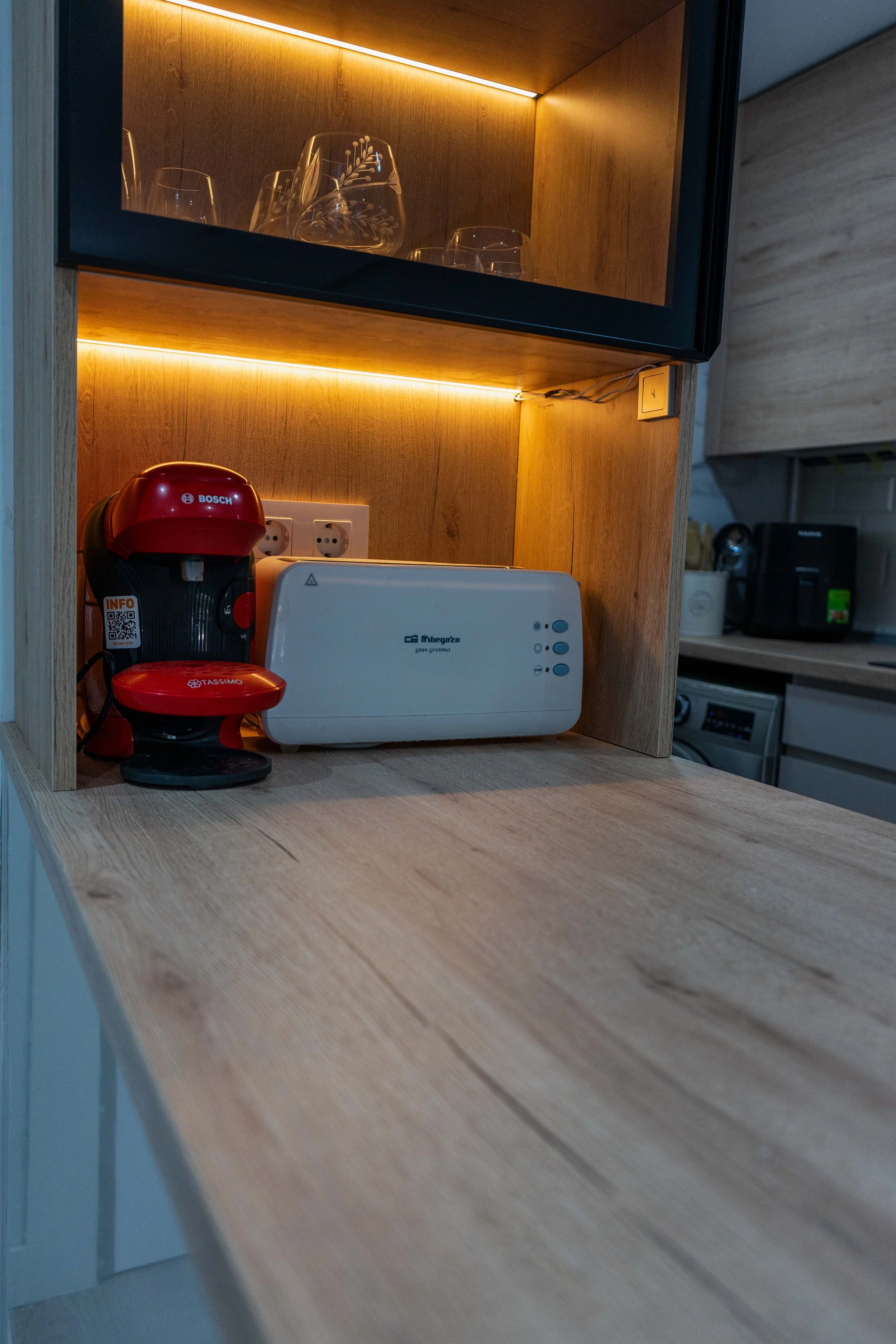 Una repisa de madera con vasos de cristal decorados, una máquina de café portátil roja y una caja de limpieza de Lavazza en una cocina moderna con electrodomésticos.