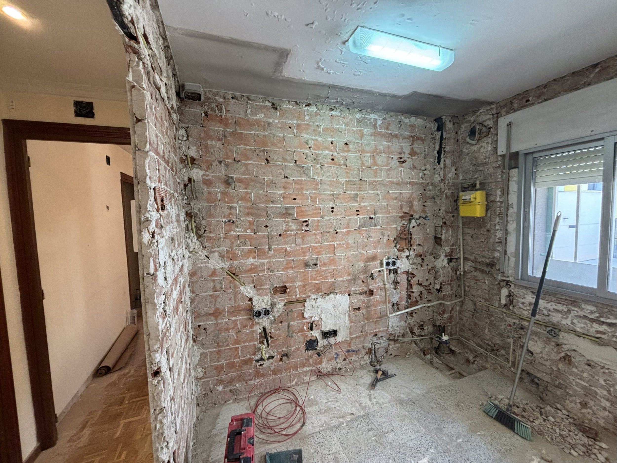 Una habitación en construcción con paredes de ladrillo expuesto y cables. Hay una ventana, una escoba y herramienta en el suelo, y una luz fluorescente en el techo.