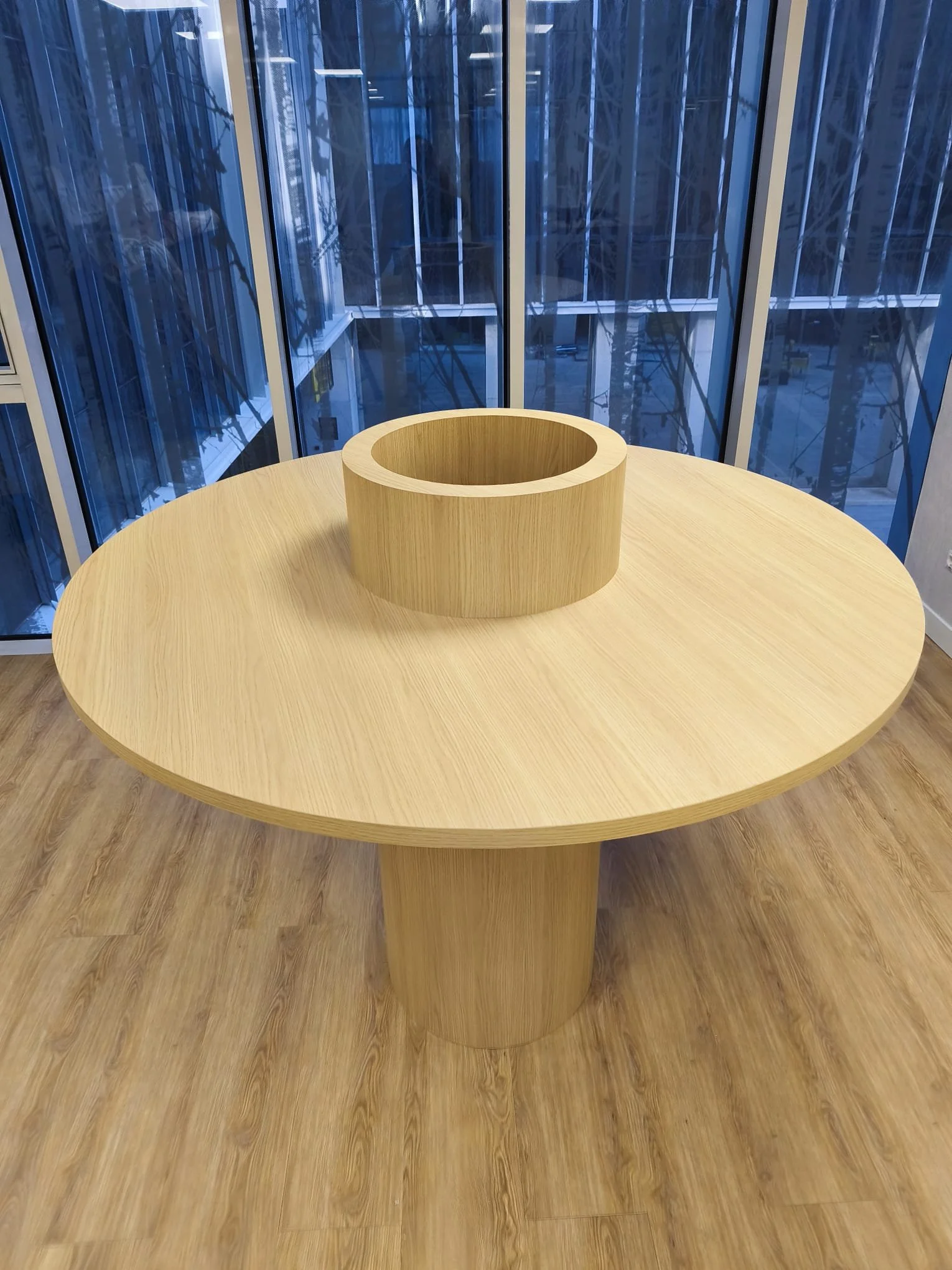 Mesa redonda de madera clara con un adorno circular en el centro, en una oficina con ventanas de vidrio.