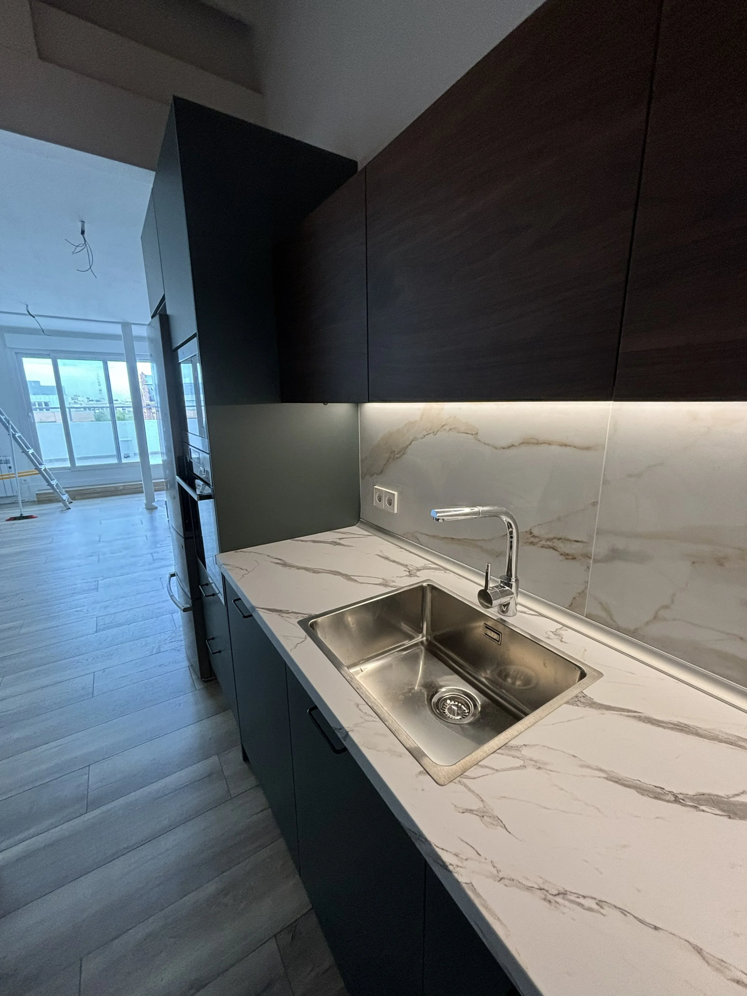 Cocina moderna con encimera de mármol, fregadero de acero inoxidable y gabinetes oscuros. Ventana grande en fondo con vista a una sala en construcción.