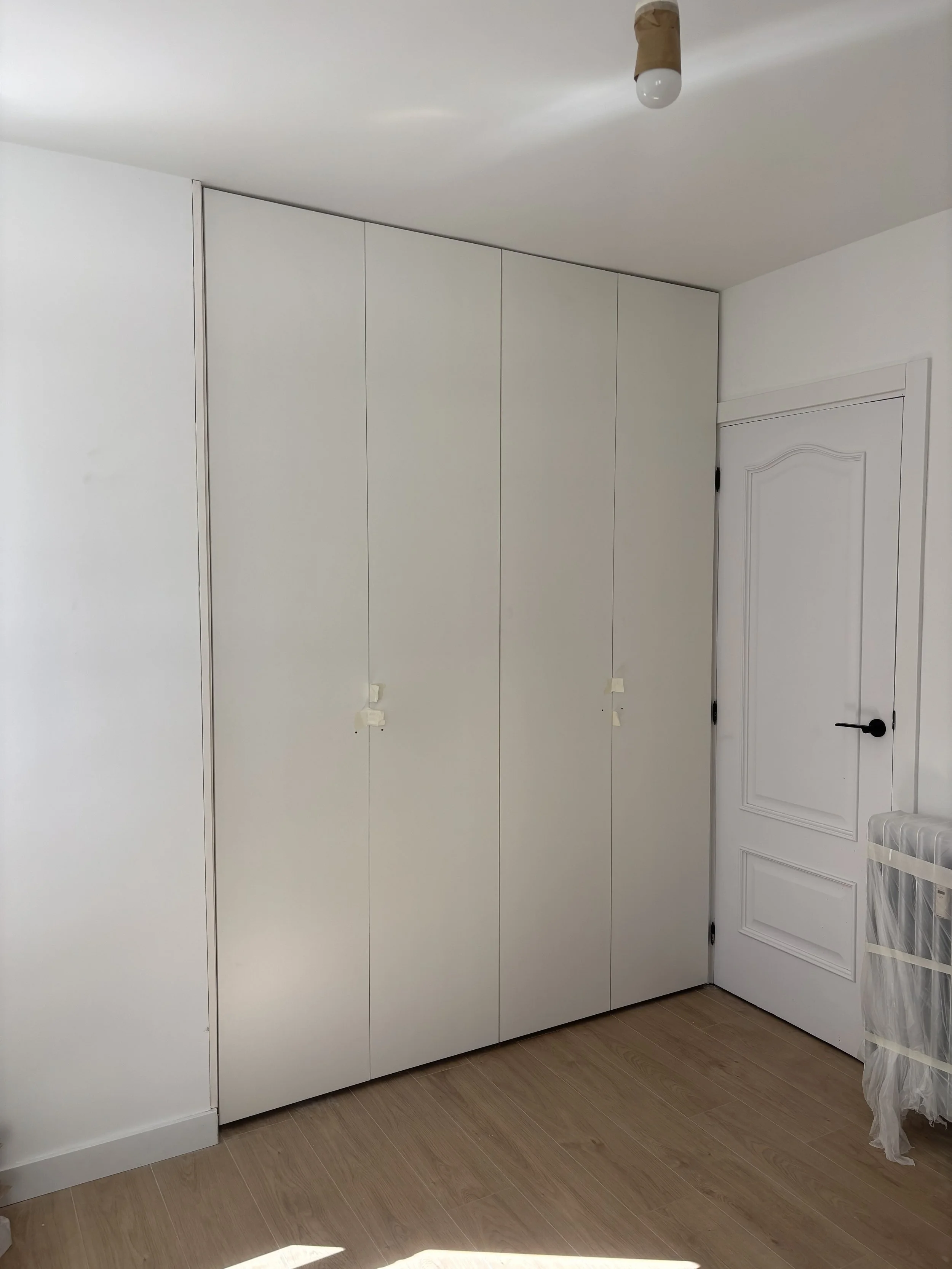 Armario blanco empotrado en una habitación, cerca de una puerta blanca con mango negro, y una caja de plástico cubierto con tela en el lado derecho.