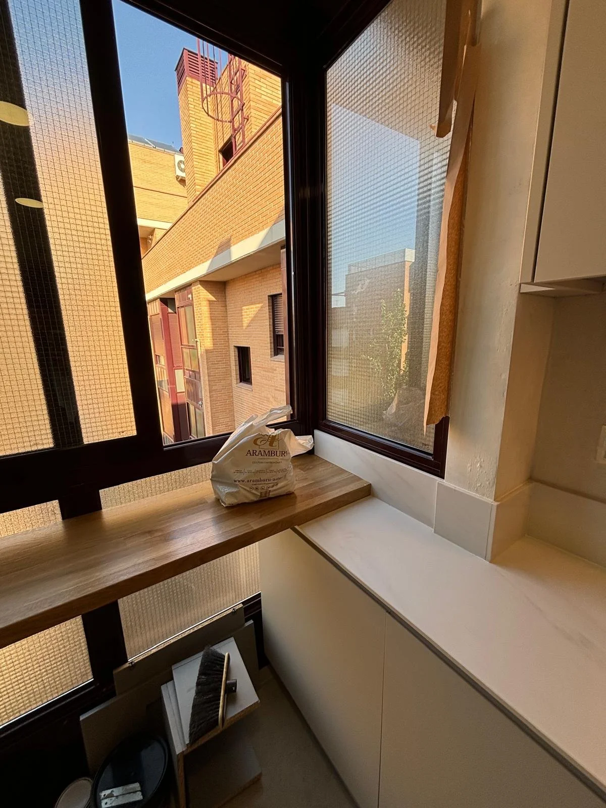 Vista desde una ventana de un apartamento con una bolsa con la marca Aramburí en el alféizar y utensilios de limpieza—una escobilla y una bandeja—sobre una mesa de madera pequeña.