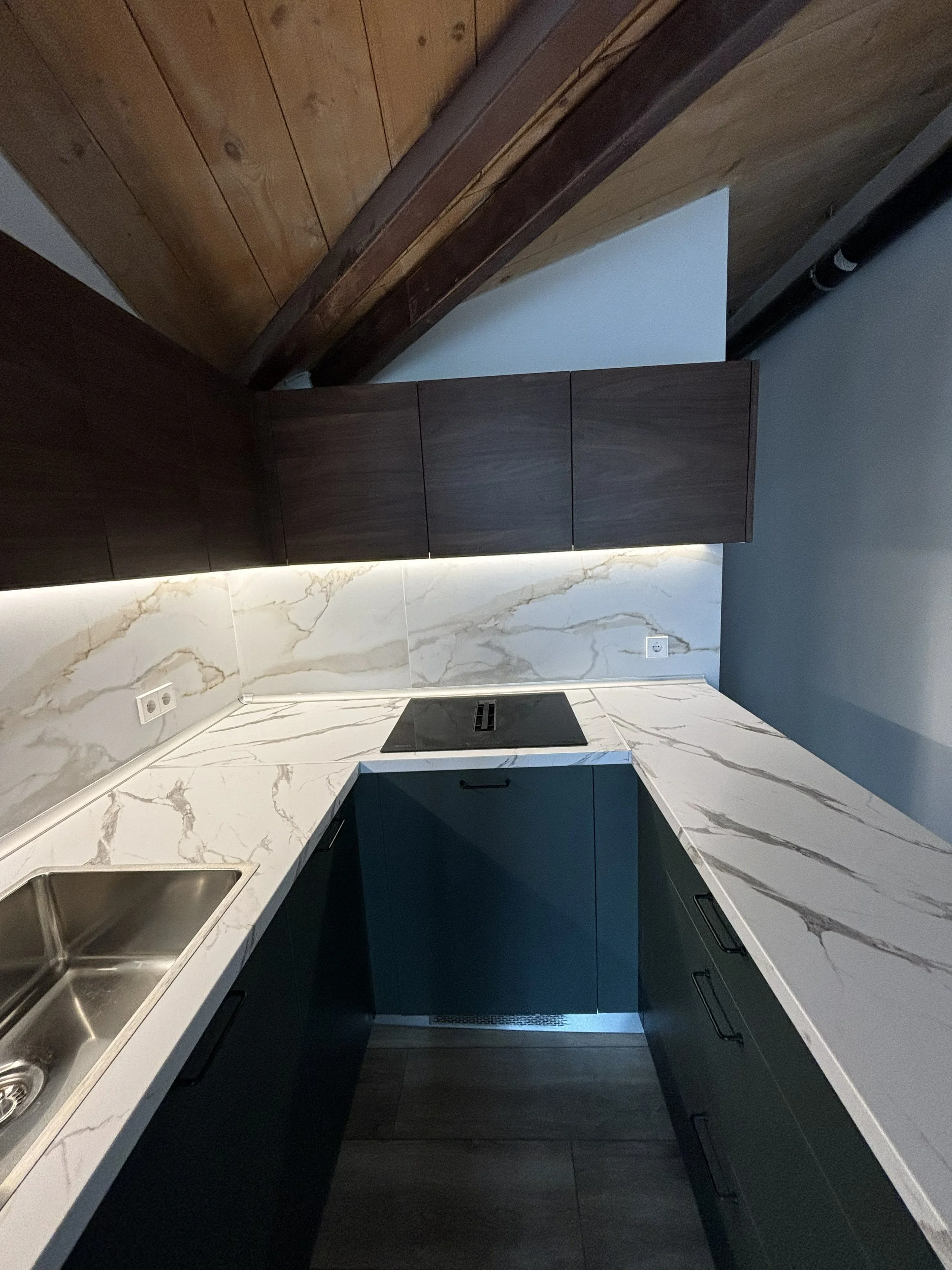 Cocina moderna con encimera de mármol, gabinete azul oscuro y paredes de cerámica con vetas. Inodoro oculto y ventanales con techo de madera.