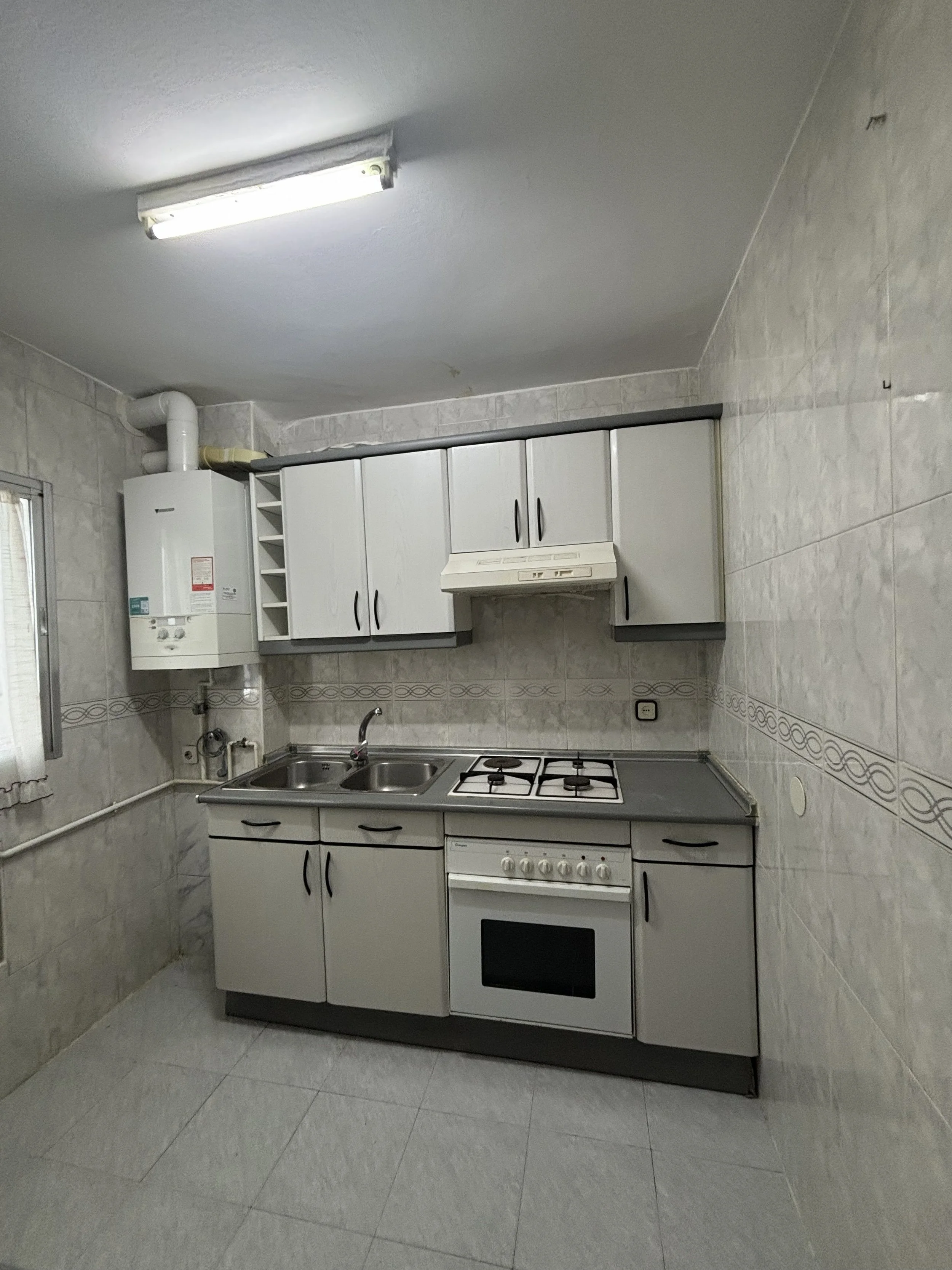 Pequeña cocina con gabinetes blancos, estufa de cuatro quemadores, fregadero doble y paredes de azulejos claras