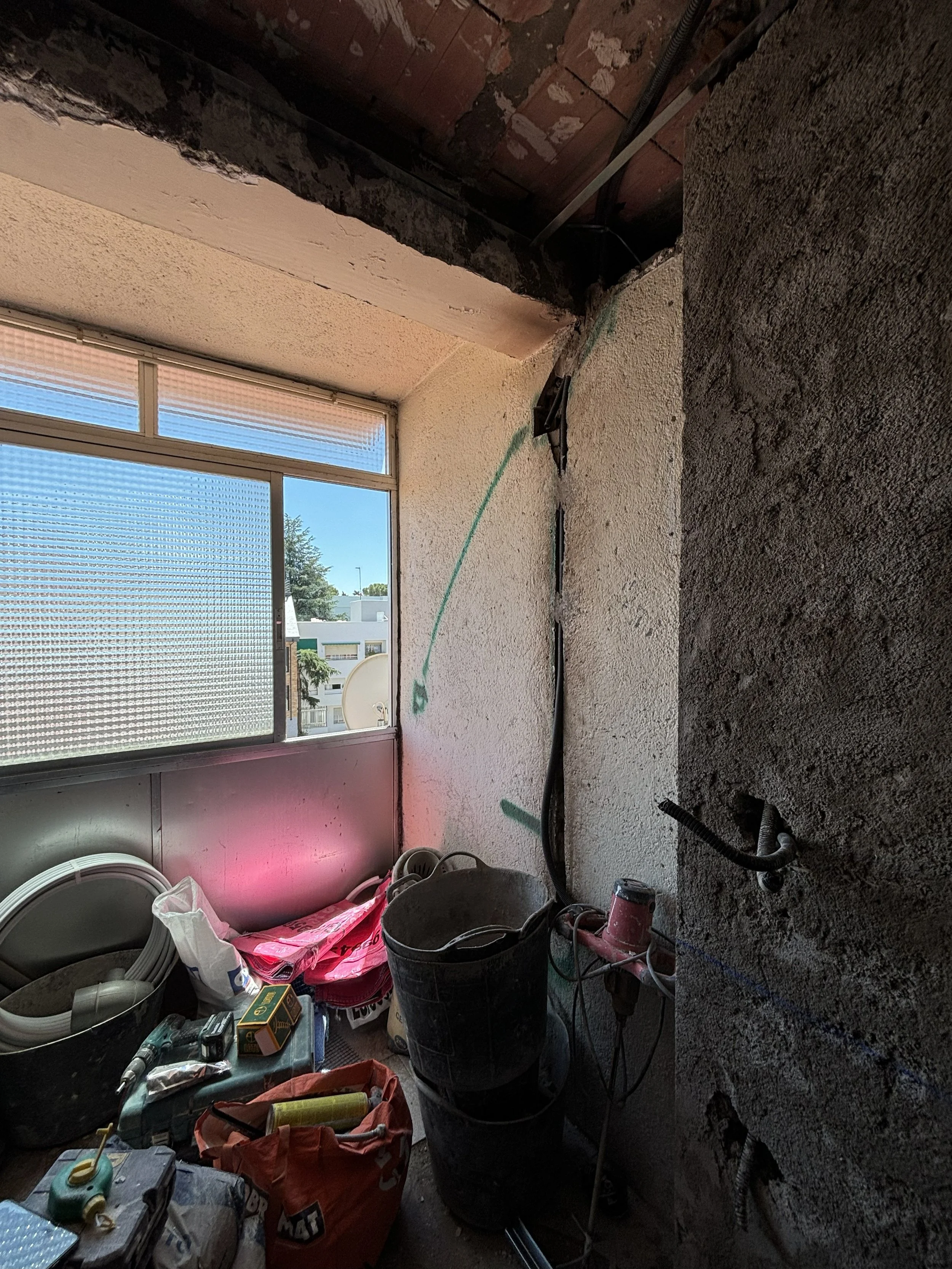 Espacio de construcción o renovación con herramientas, cables, cubetas y materiales en una habitación con ventanas, paredes en proceso de reparación y un techo parcialmente abierto.