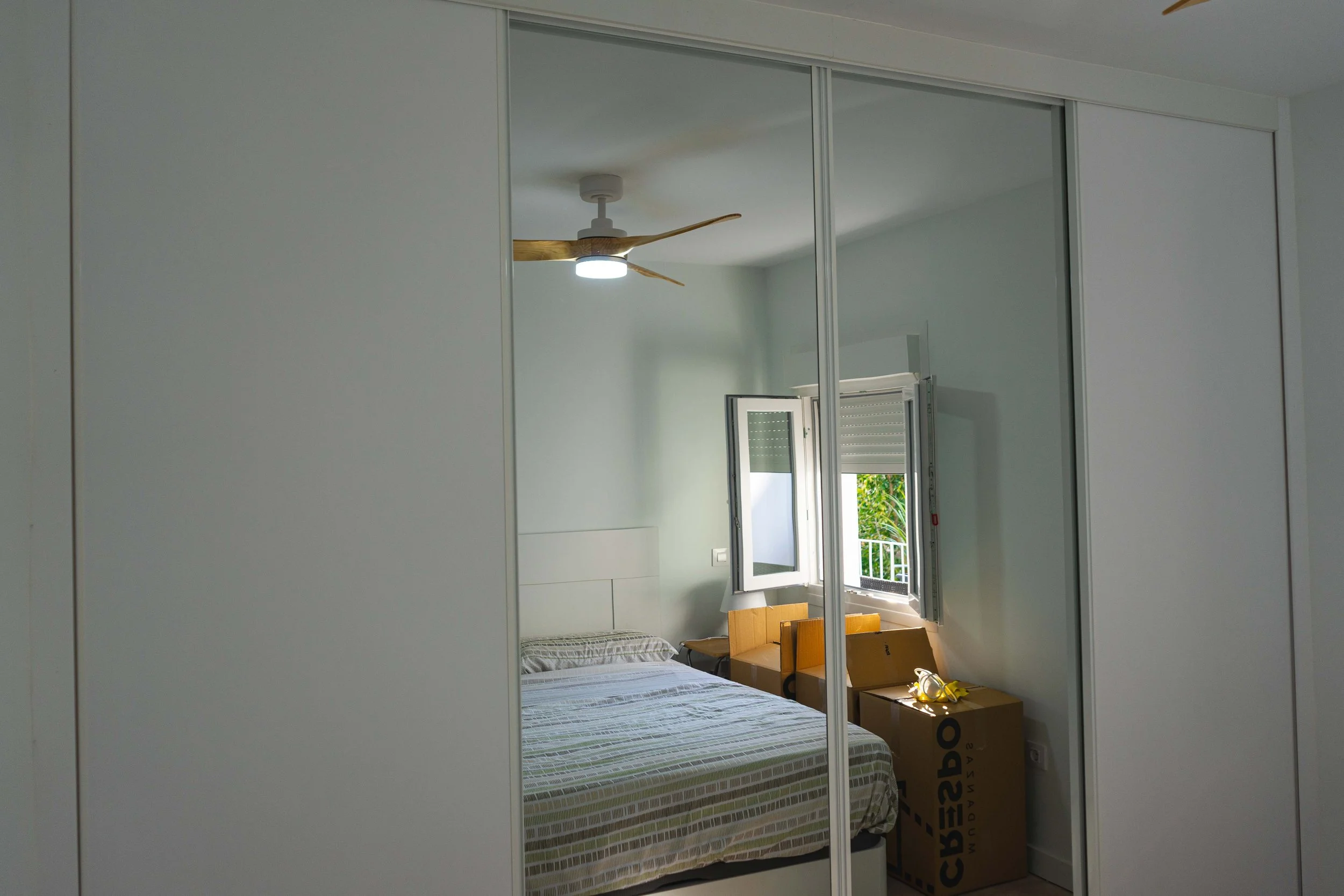 Habitación con armario de puertas corredizas, cama, ventanas abiertas con vista a árboles, ventilador de techo y cajas de mudanza en el suelo.