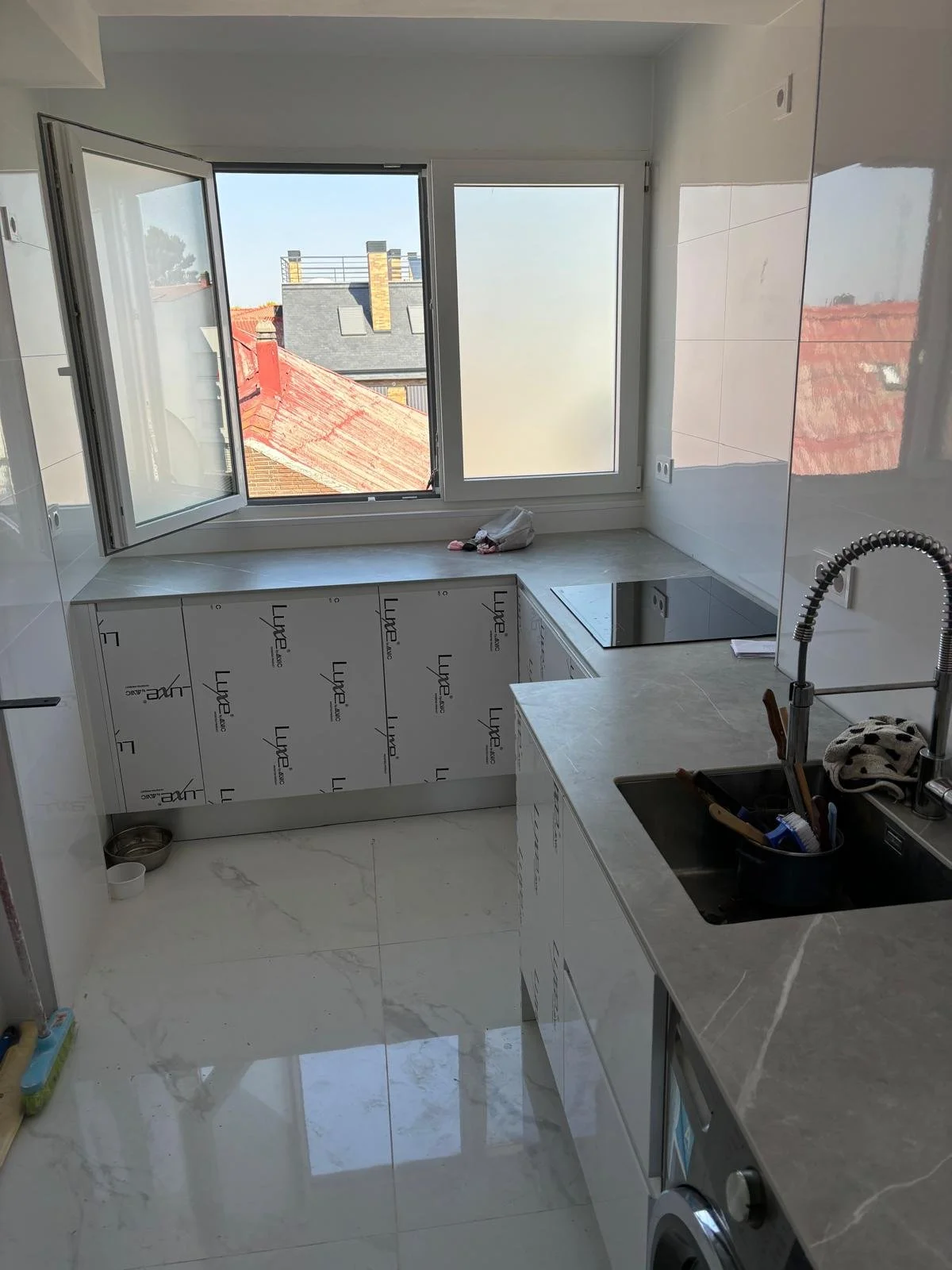 Cocina moderna con ventanas grandes, encimera de piedra, fregadero con grifo flexible, y maquinaria instalada, con baldas y utensilios de cocina al lado del fregadero.