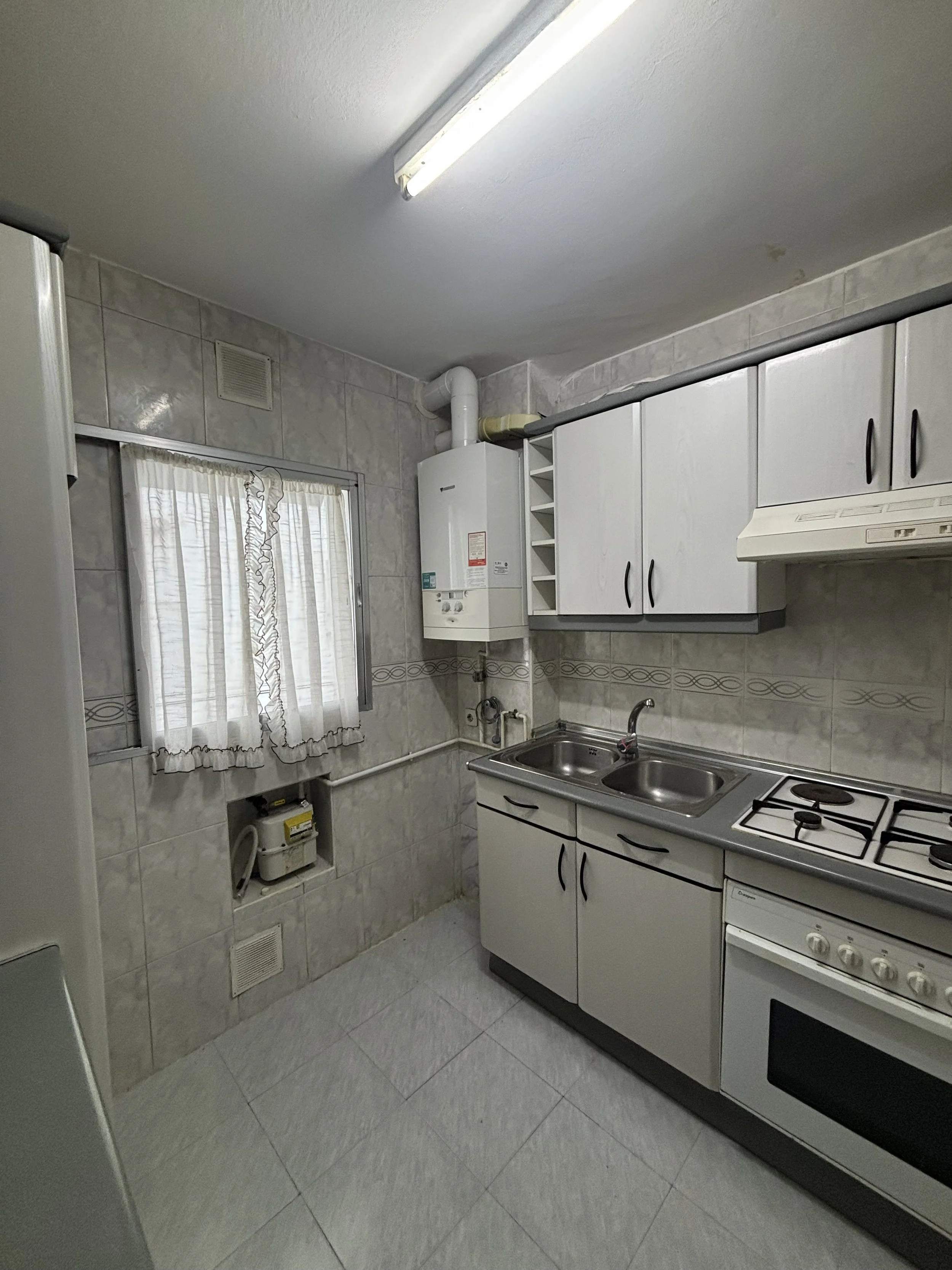 Cocina pequeña con gabinetes blancos, estufa y fregadero, ventanales con cortinas y paredes de azulejos, un calentador de agua en la pared.