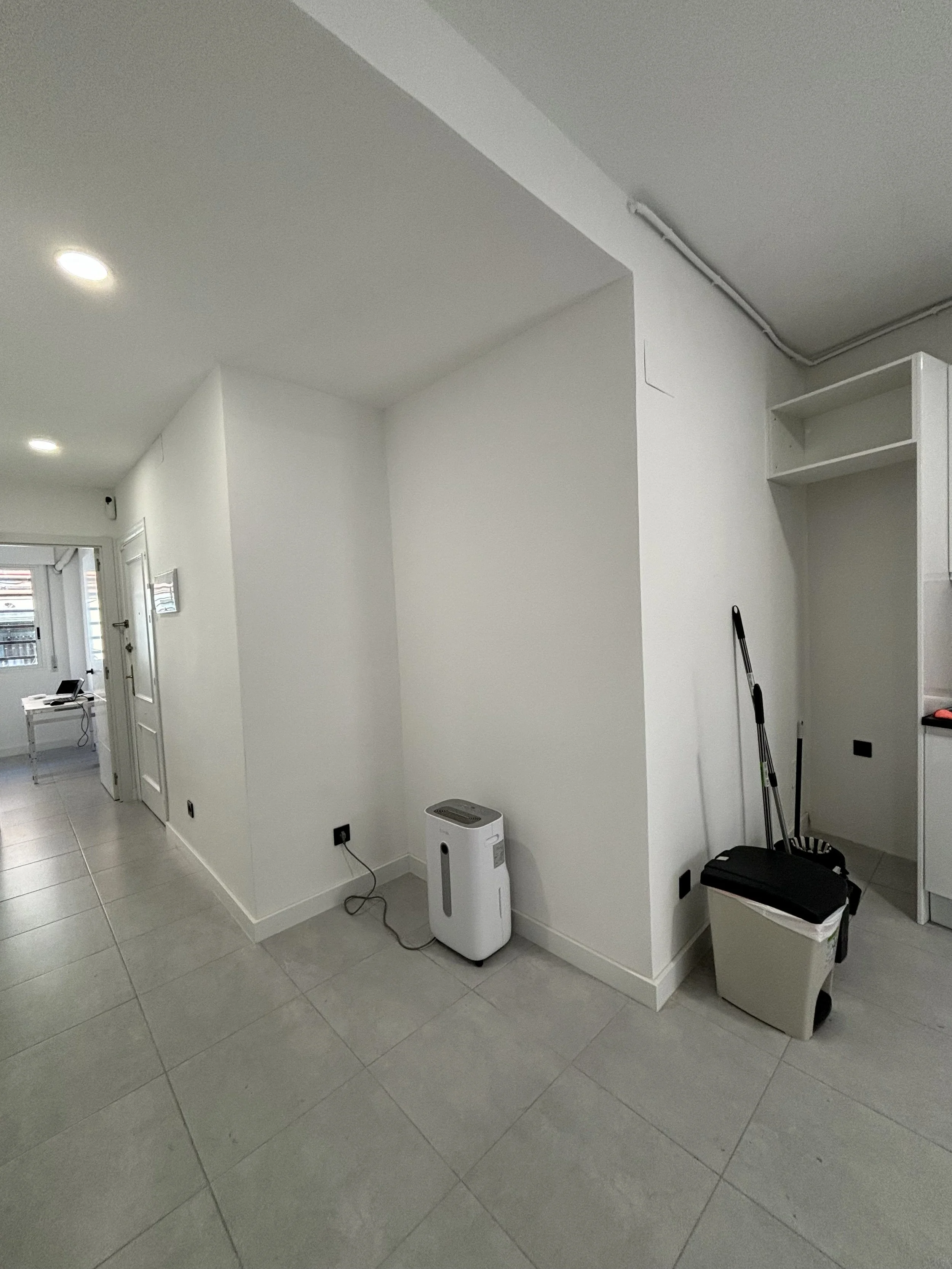 Interiores de una oficina o área de trabajo con paredes blancas, suelo de baldosas grises, en el fondo una mesa con teléfono y sillas, en primer plano un deshumificador y en el rincón algunas herramientas y una caja con objetos.